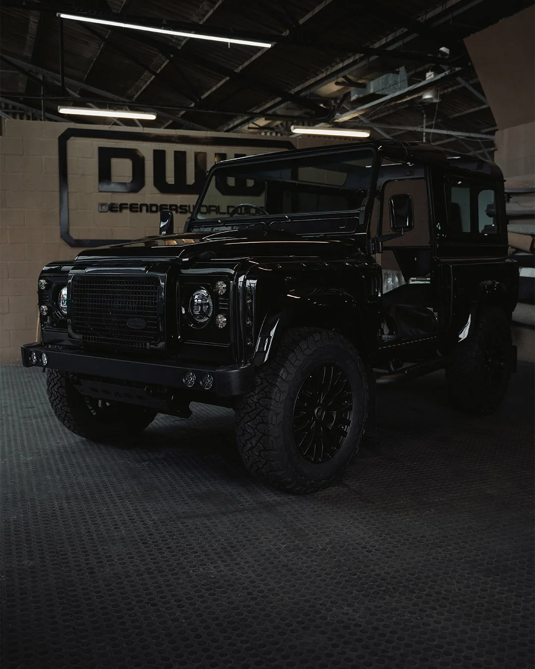 A black Land Rover Defender inside a workshop with a sign that says Defenders Worldwide.