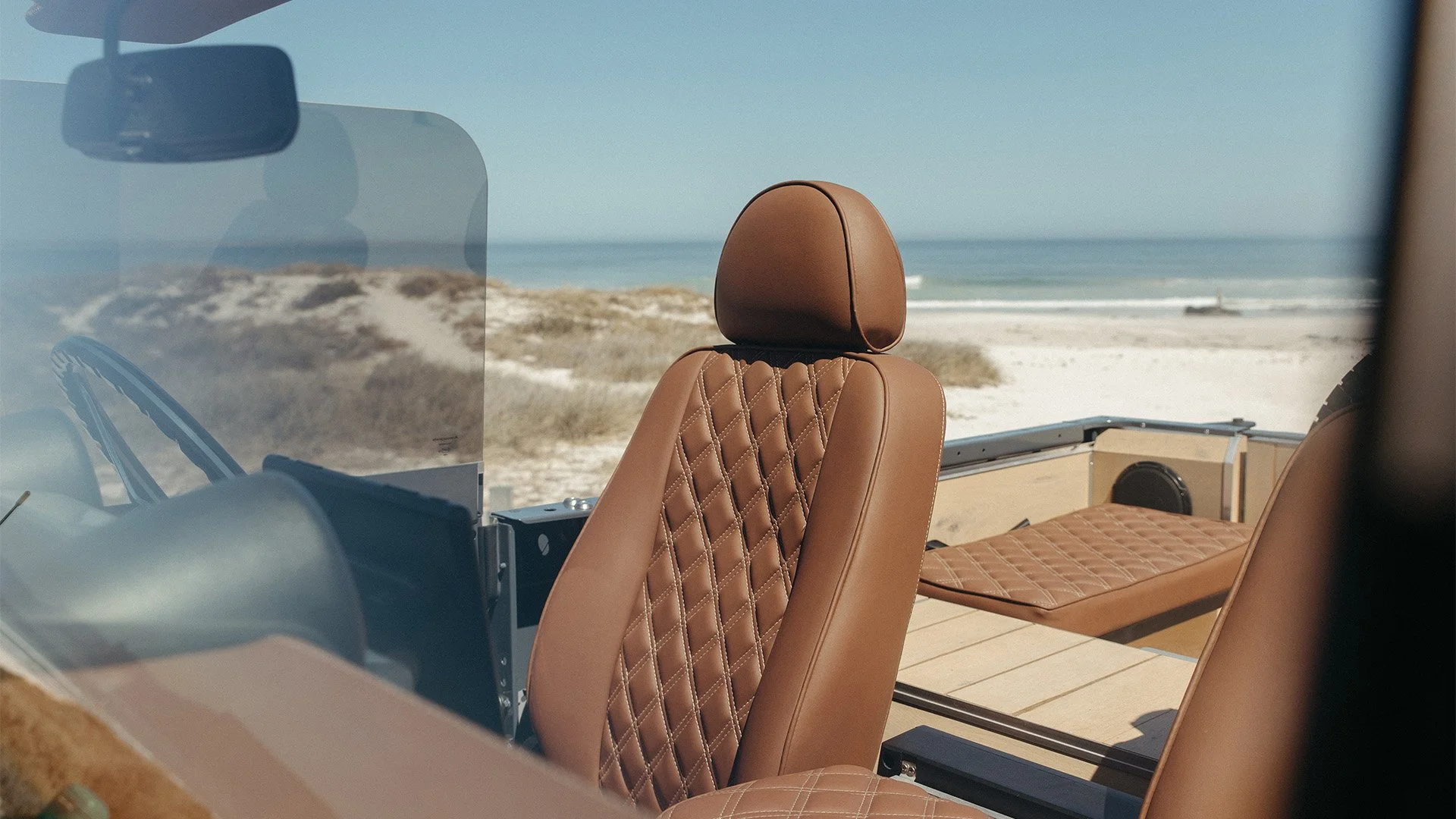 Interior view of a vehicle with tan quilted leather seats, overlooking a beach with sand dunes and the ocean in the background.