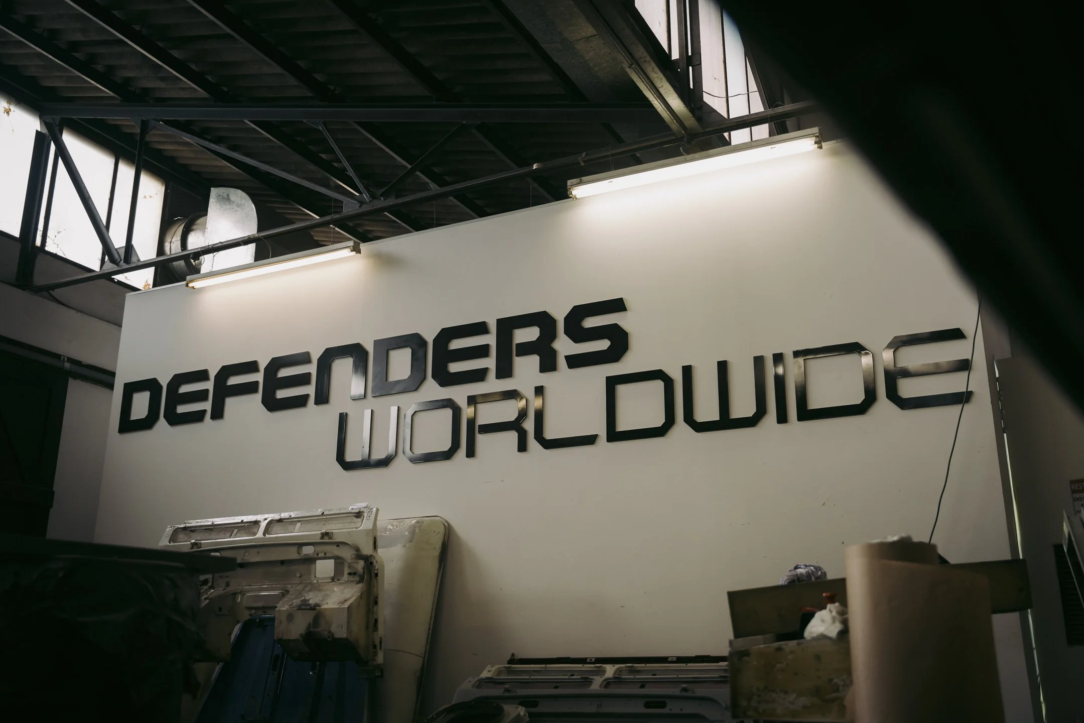 View of an industrial interior with a white wall displaying black 3D letters spelling 'DEFENDERS' and 'WORLD' underneath. The ceiling has exposed black metal beams and pipes, with fluorescent lights. An equipment or machine is partially visible in th