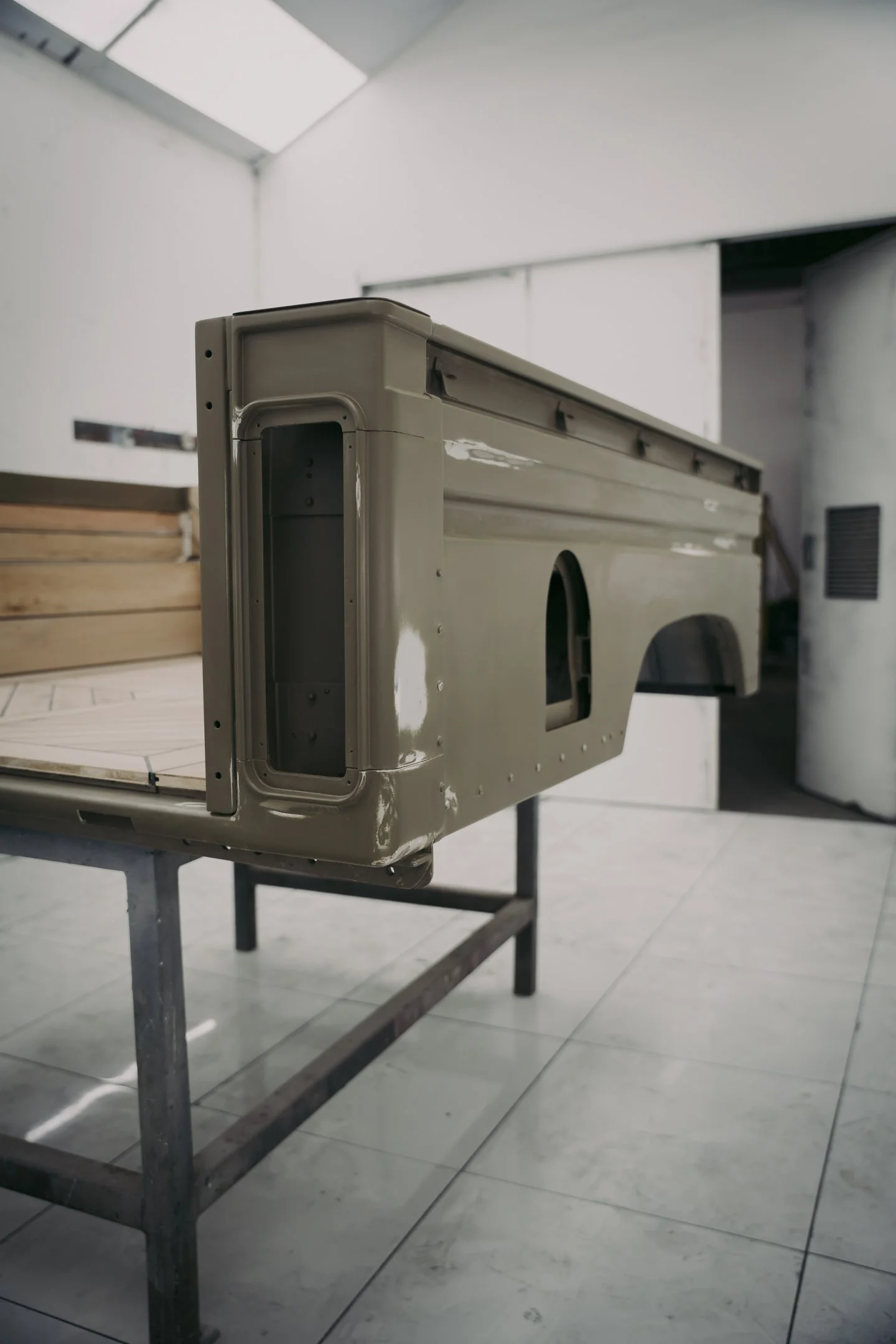 A partially assembled vintage style Land Rover Defender shell placed on a metal work table in an indoor space with tiled flooring and white walls.