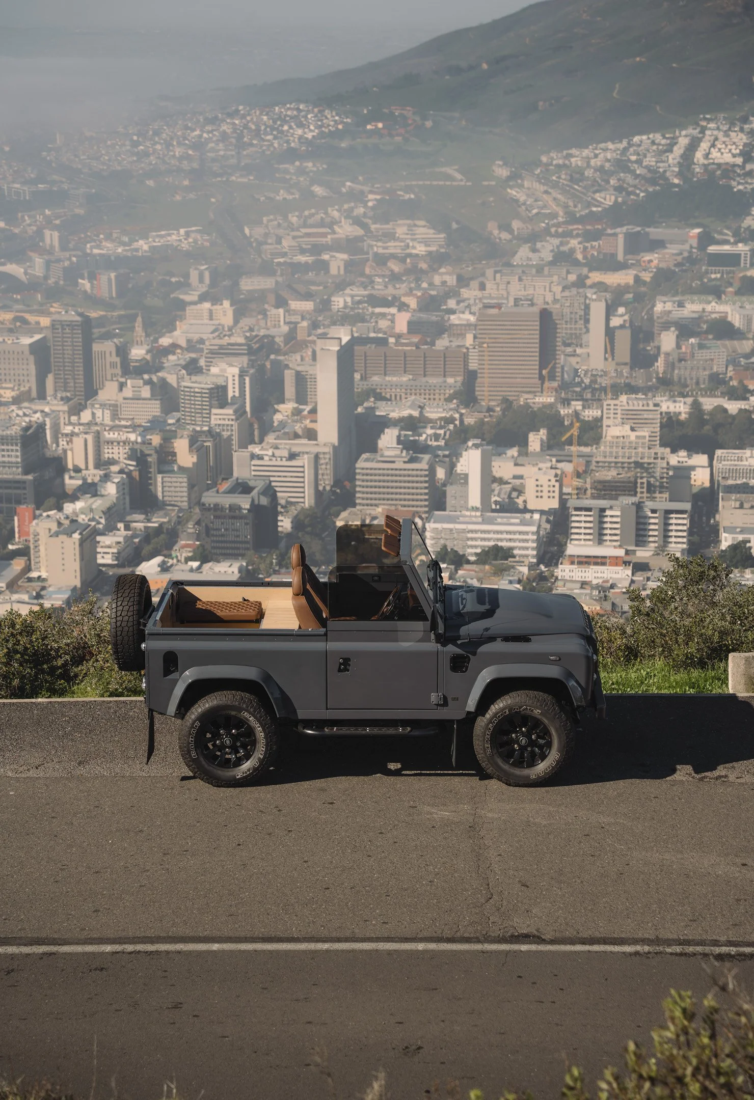 A black off-road Land Rover Defender parked on a road with a cityscape and hills in the background.