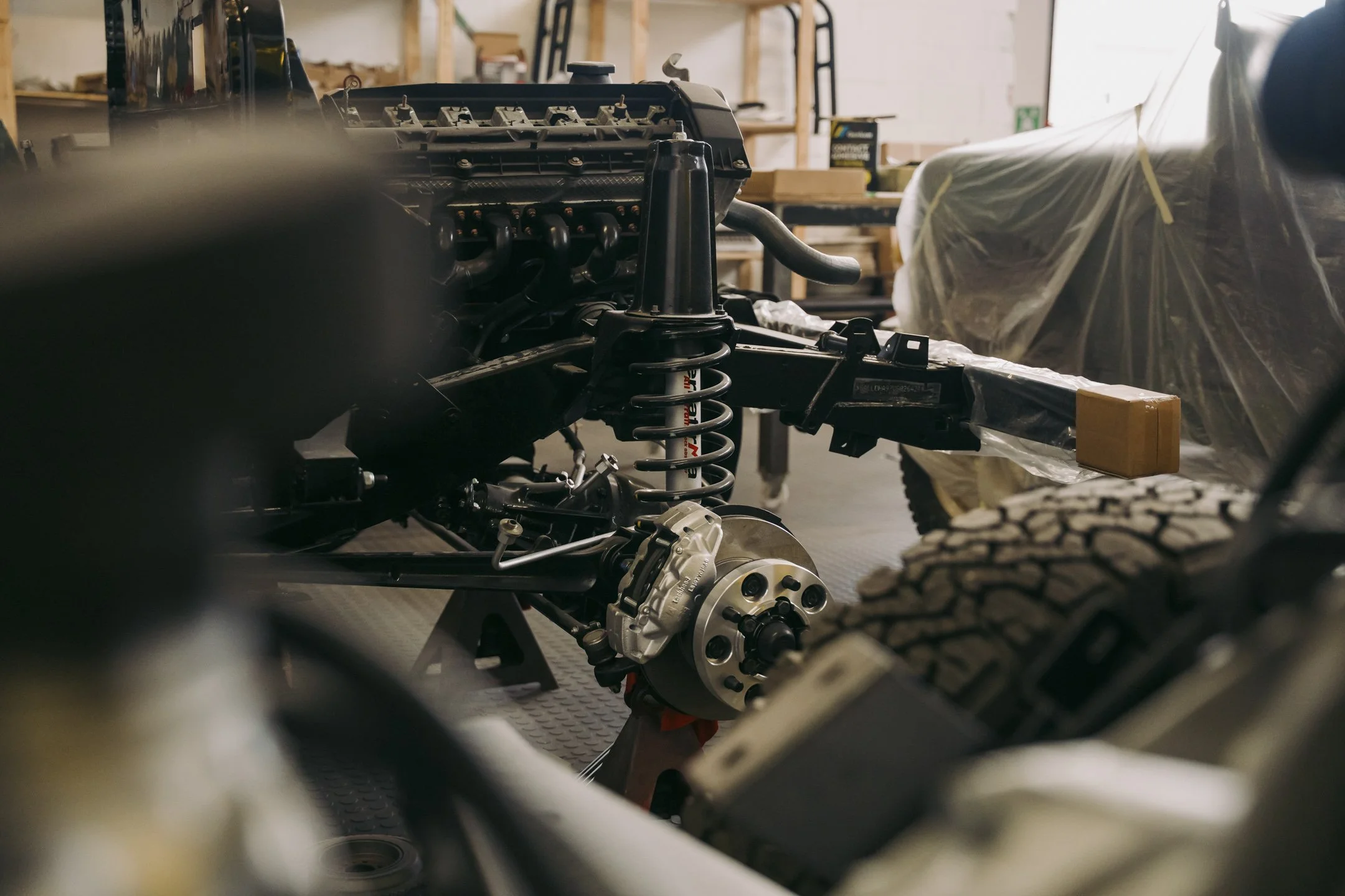 Partially assembled vehicle chassis in a workshop, showing suspension components, an engine, and surrounding tools and equipment.