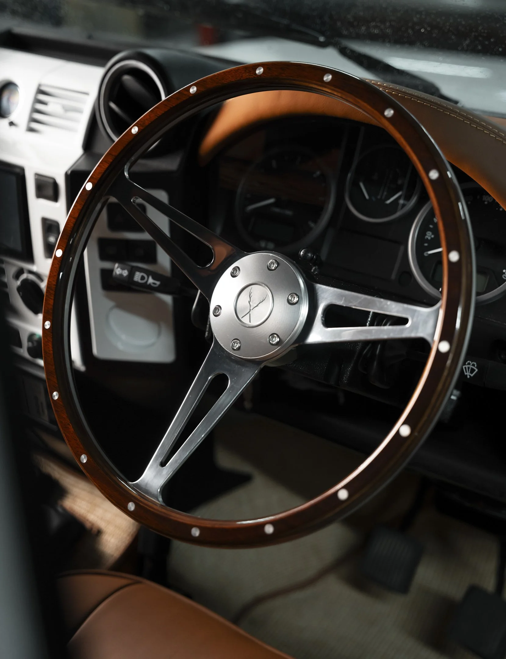 Close-up of a Land Rover Defender's interior steering wheel with a wood rim and metal spokes, dashboard gauges in the background.