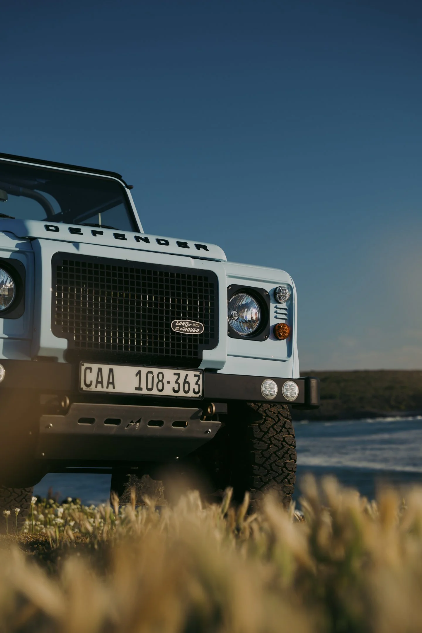 Close-up of a white Land Rover Defender off-road vehicle parked on a grassy area near water, with a clear blue sky in the background.