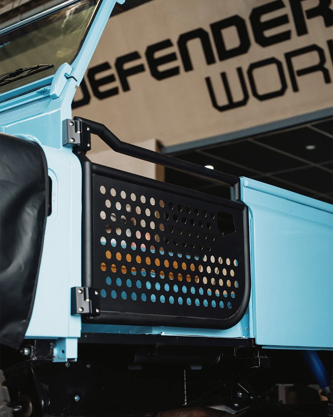 Close-up of a light blue utility vehicle with a black perforated side panel and black handrails, displayed indoors with a sign that reads 'REFERENDUM WORK' in the background.