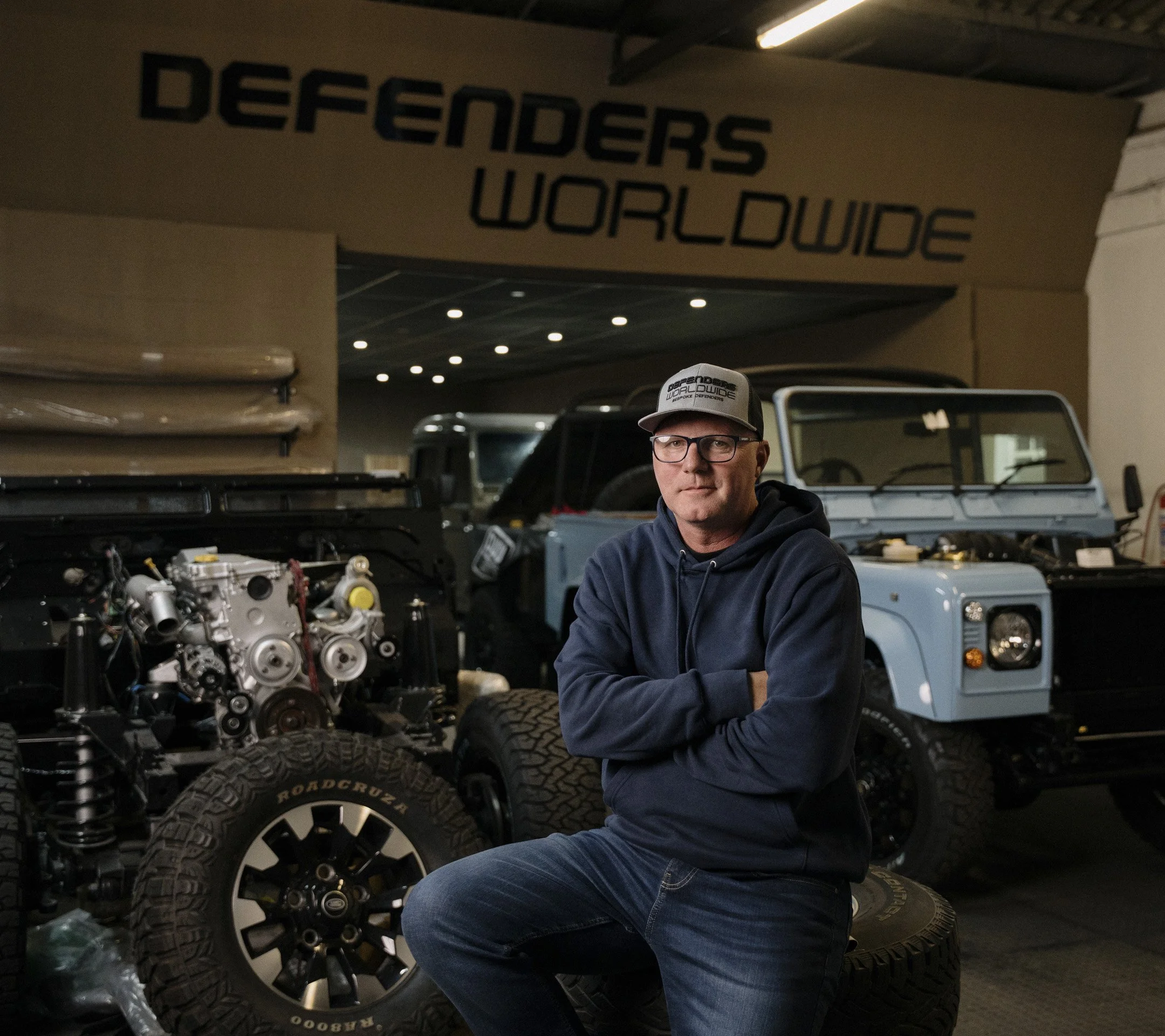 A man wearing glasses and a cap with the logo 'Defenders WorldWide' is standing in a garage or workshop surrounded by off-road tires, vehicles, and engine components, with a sign that reads 'Defenders Worldwide' in the background.