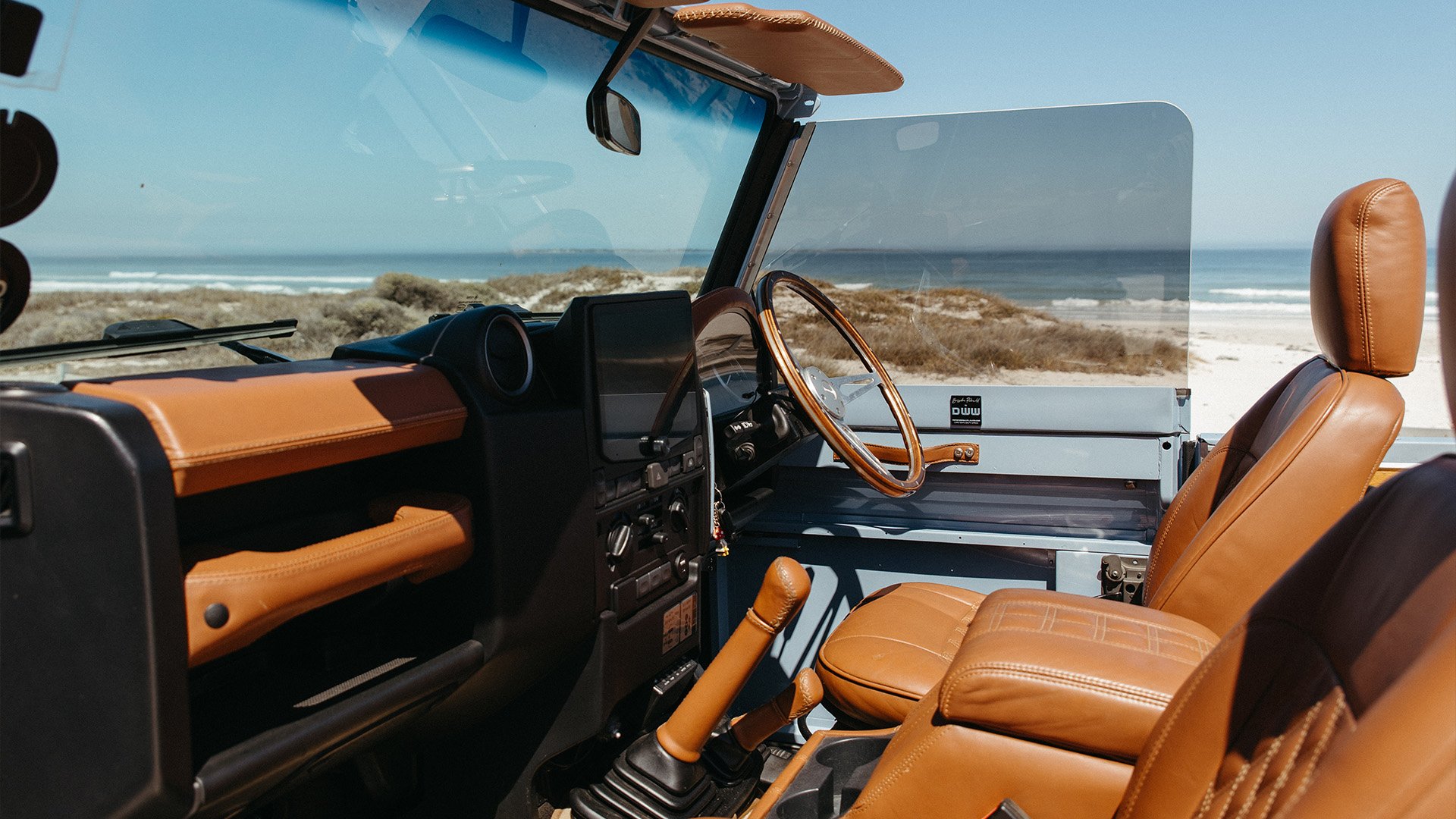 Interior of a vintage off-road vehicle parked by the beach with tan leather seats, a wooden steering wheel, a large windshield, and a view of the ocean and sand dunes in the background.