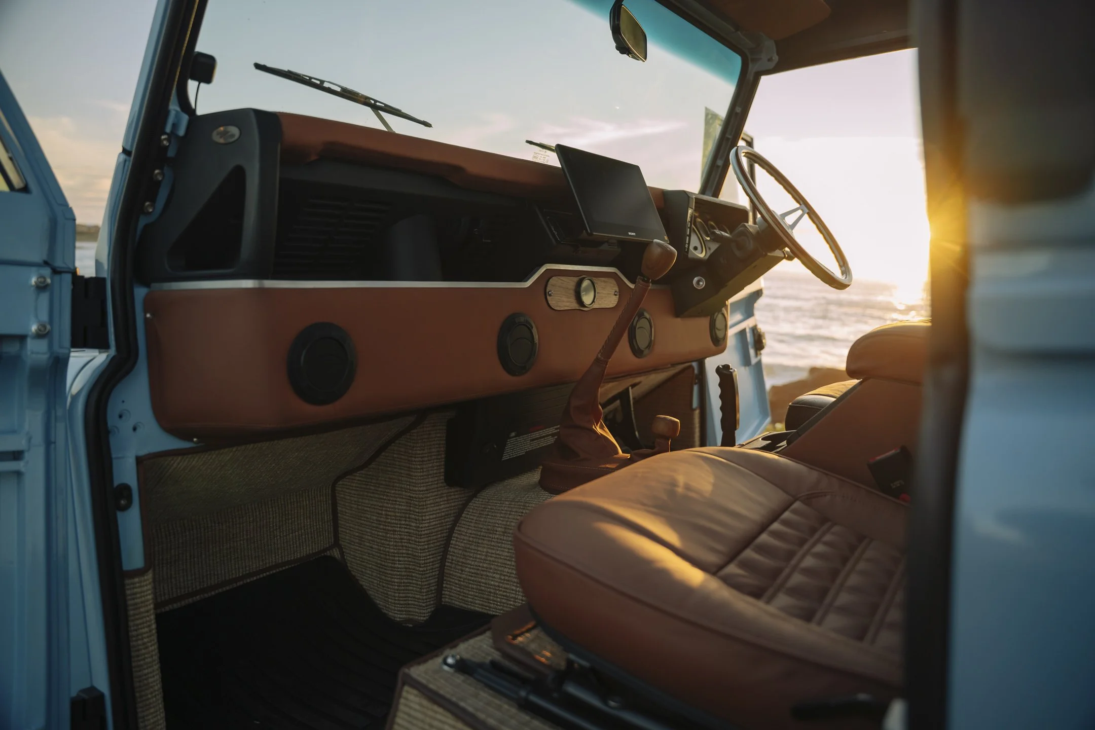 Interior of a vintage vehicle showing a dashboard, steering wheel, and beige leather seat near large windows with sunset view over water.