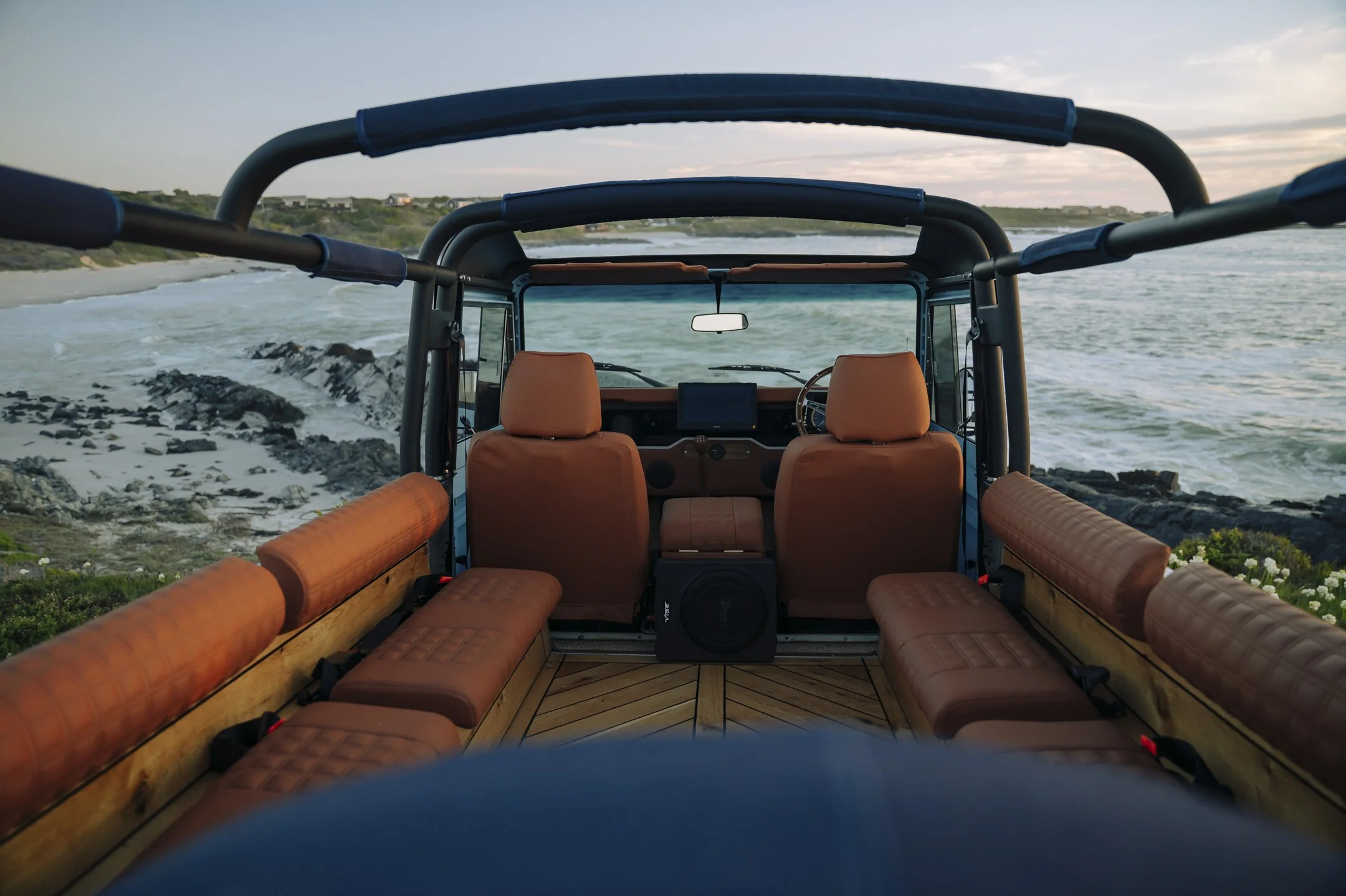 The image shows the interior of an off-road vehicle with cushioned seats, overlooking a coastline with waves crashing onto rocks and a beach, under a partly cloudy sky.