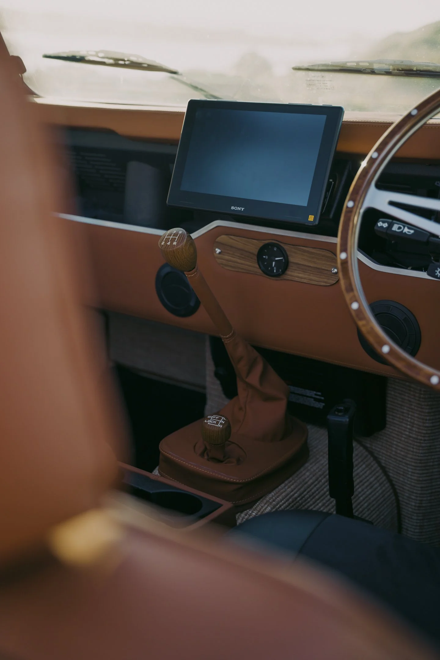 Interior of a vintage car with a wooden steering wheel, two gear shifters with wooden knobs, a Sony screen mounted on the dashboard, and a small clock, with a foggy windshield showing a blurred landscape outside.