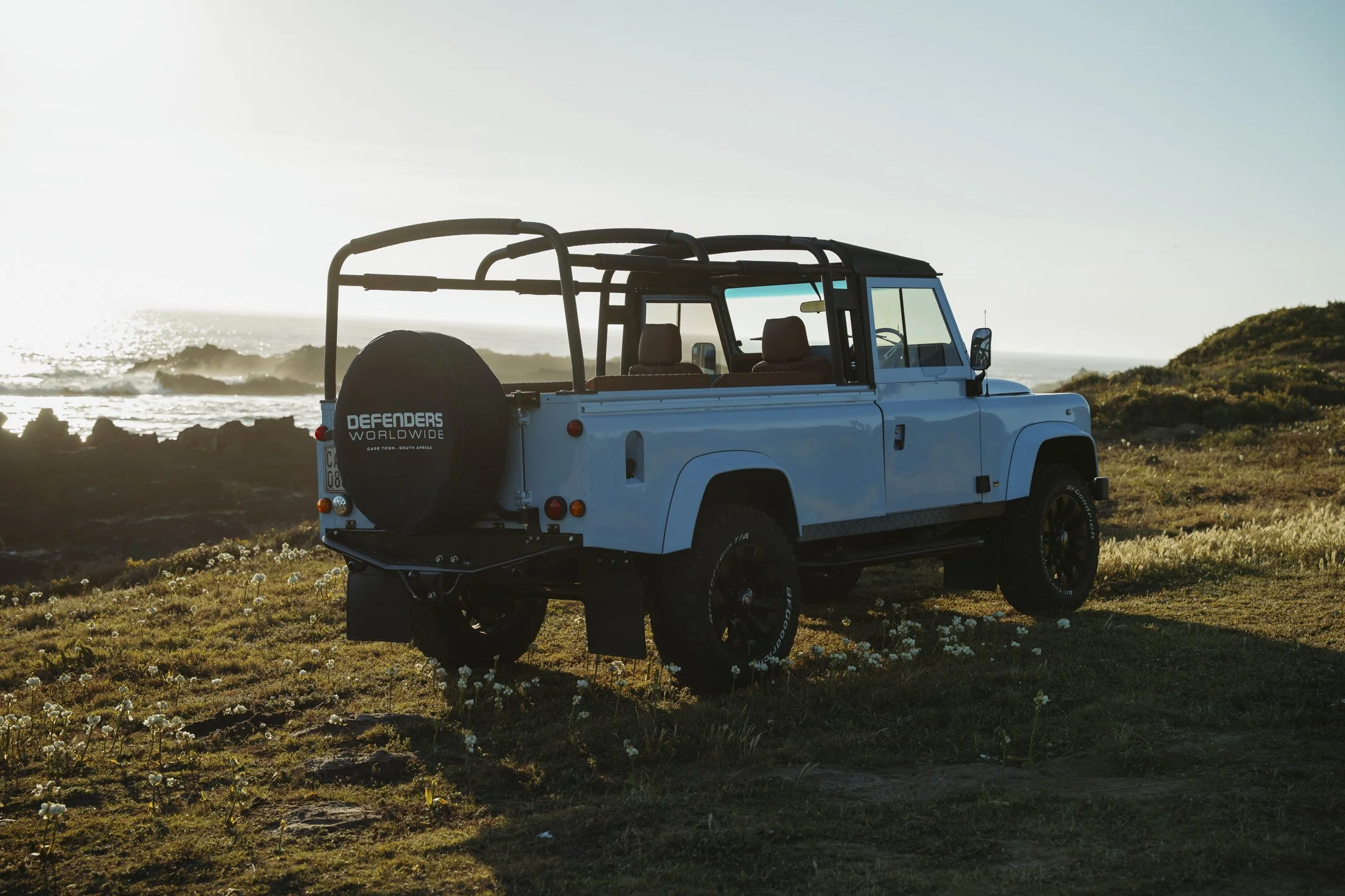 A blue off-road vehicle parked on grassy terrain near the coast with the ocean and rocks in the background, sunlight illuminating the scene.