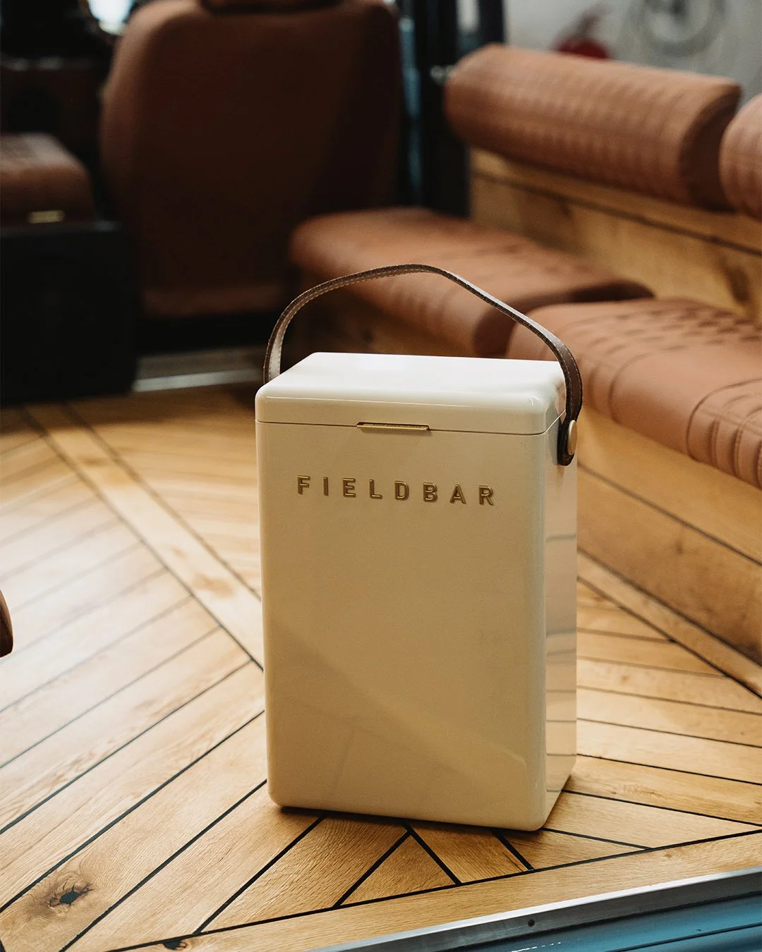 A white rectangular cooler with a leather strap handle and gold lettering that reads 'FIELDBAR' on the front, placed on a wooden table in a cozy indoor setting with brown upholstered chairs and a wooden floor.