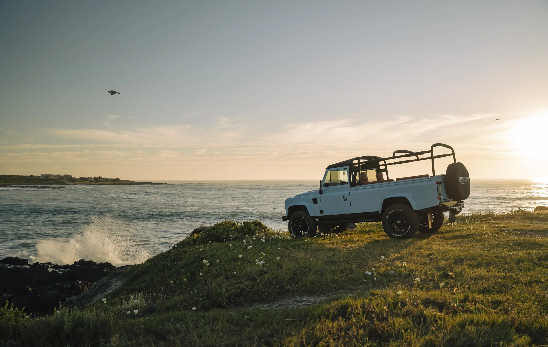 A blue off-road vehicle parked on a grassy hill near the coast with the ocean and a sunset in the background, two birds flying in the sky, and a few houses in the distance.