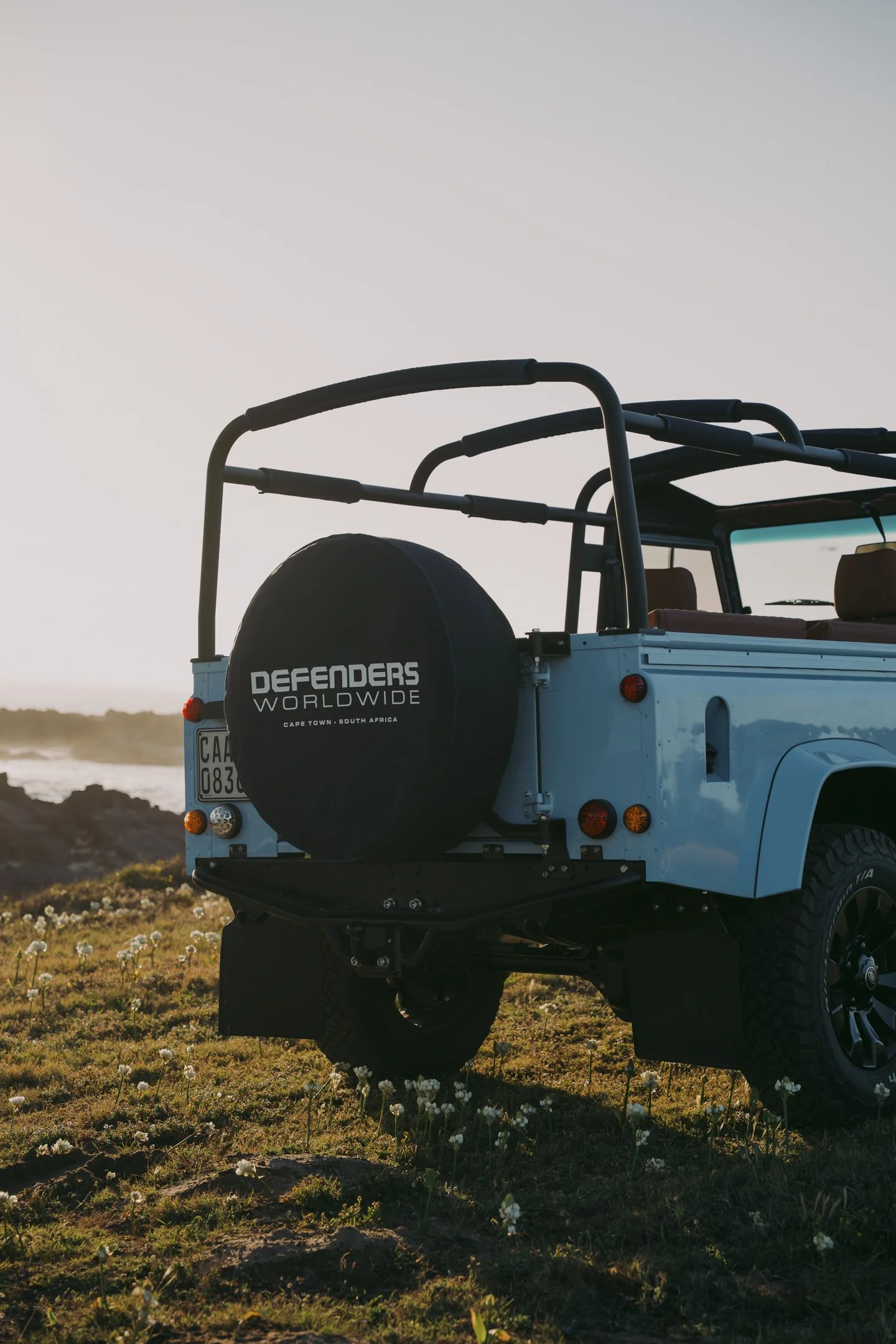 A light blue off-road vehicle with a spare tire on the back, displaying a sticker that says 'Defenders Worldwide, Cape Town, South Africa,' parked on a grassy field with small white flowers, during sunset or sunrise.