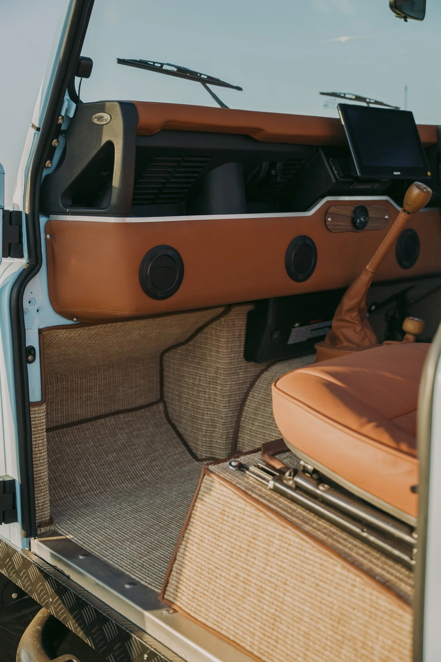Interior of a vintage Land Rover, showing the dashboard with a small digital screen, wood trim, and circular vents. The gear shift is in the foreground, wrapped in matching brown leather. The floor is carpeted in a tan checkered pattern.