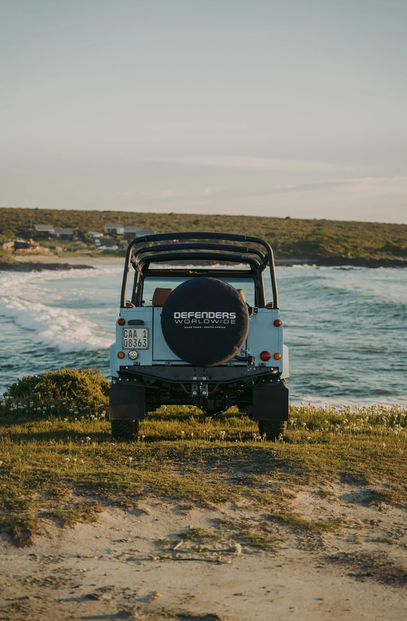 A view from behind a white off-road vehicle with a spare tire marked 'Defenders Worldwide' on a grassy coastline, facing the ocean with small waves, with a hilly landscape and a few houses in the background under a pale blue sky.