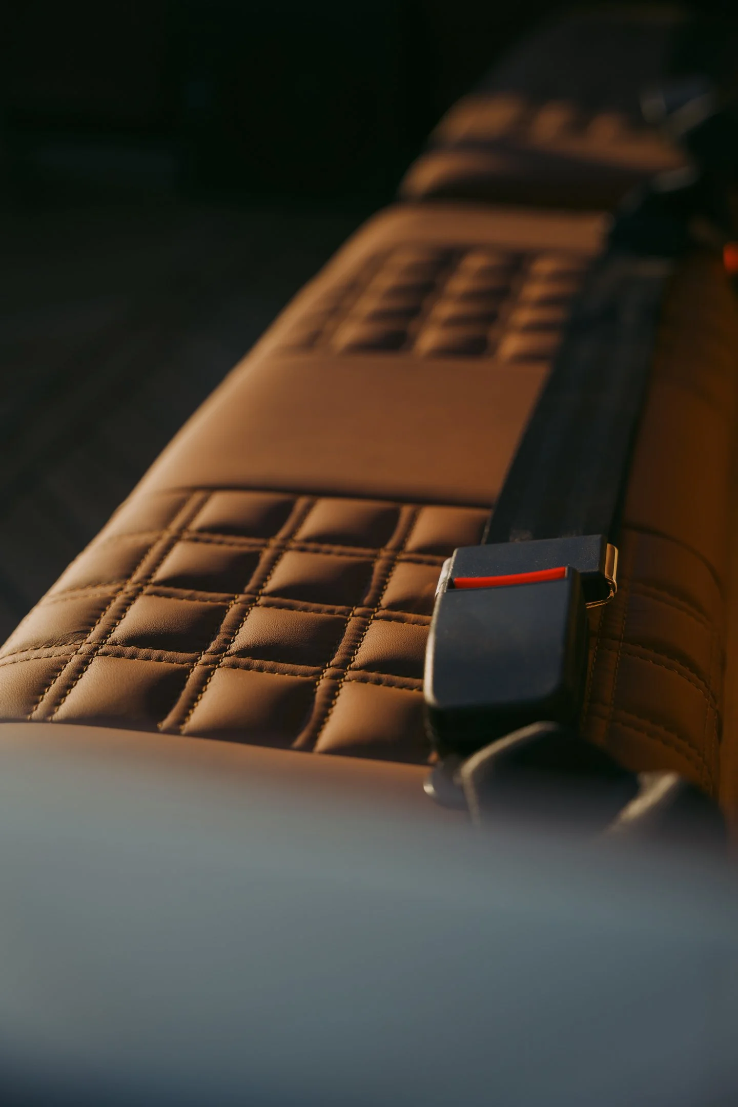 Close-up of a brown leather car seat with quilted stitching and a black seat belt buckle with a red release button.