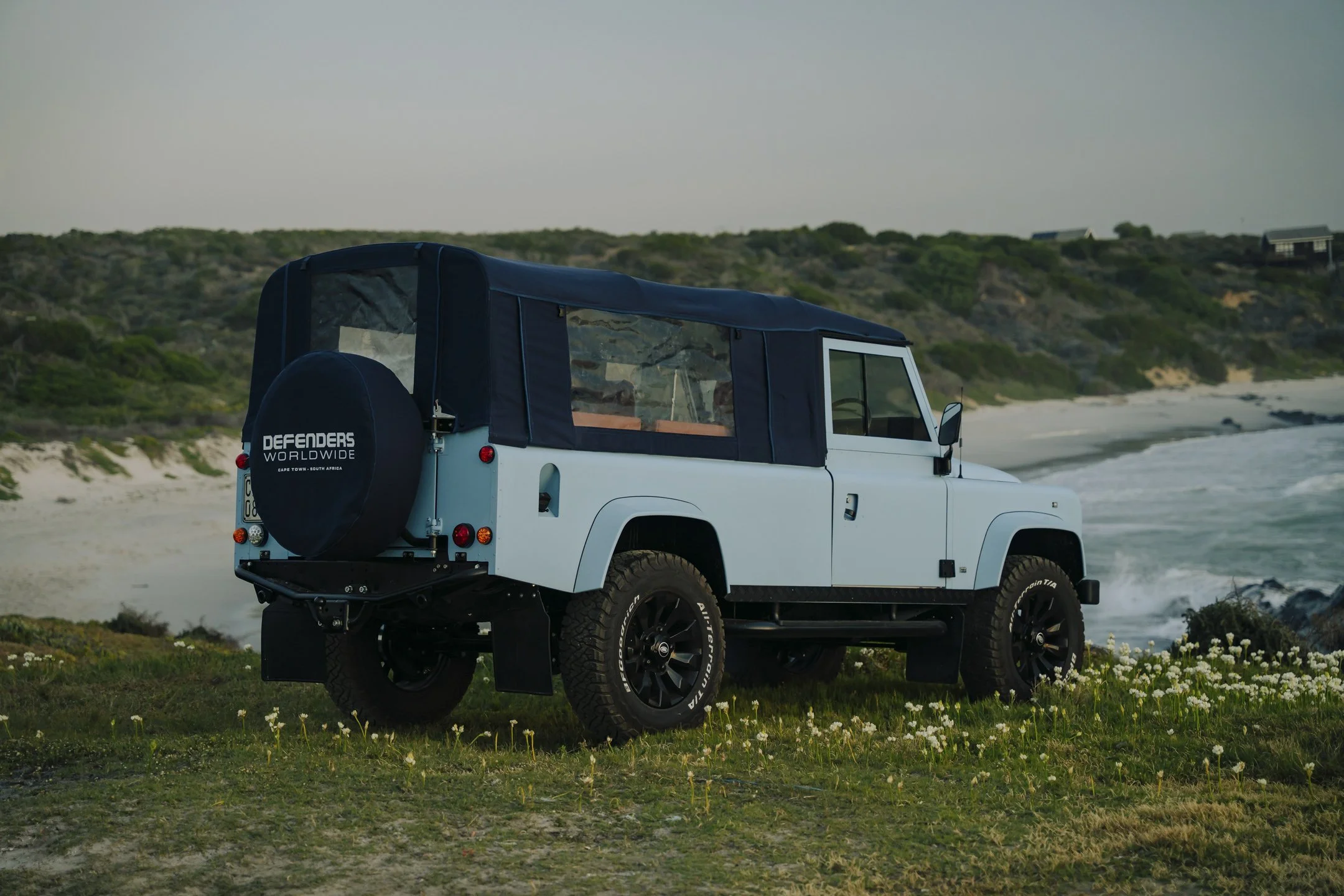 White off-road vehicle parked on a grassy area near the beach with a blue canopy and spare tire at the back, overlooking a sandy and rocky coastline with hills and sparse vegetation.