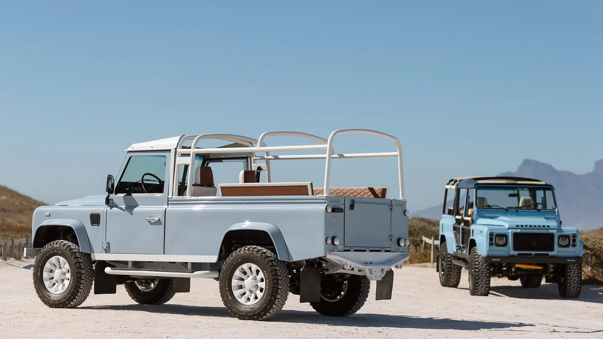 Two light blue Land Rover vehicles parked on a sandy landscape with mountains in the background, under a clear blue sky.