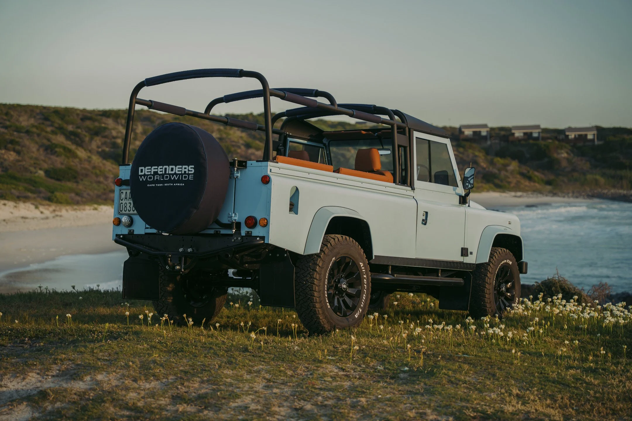 A white off-road vehicle parked on a grassy area near the beach with hills and houses in the background.