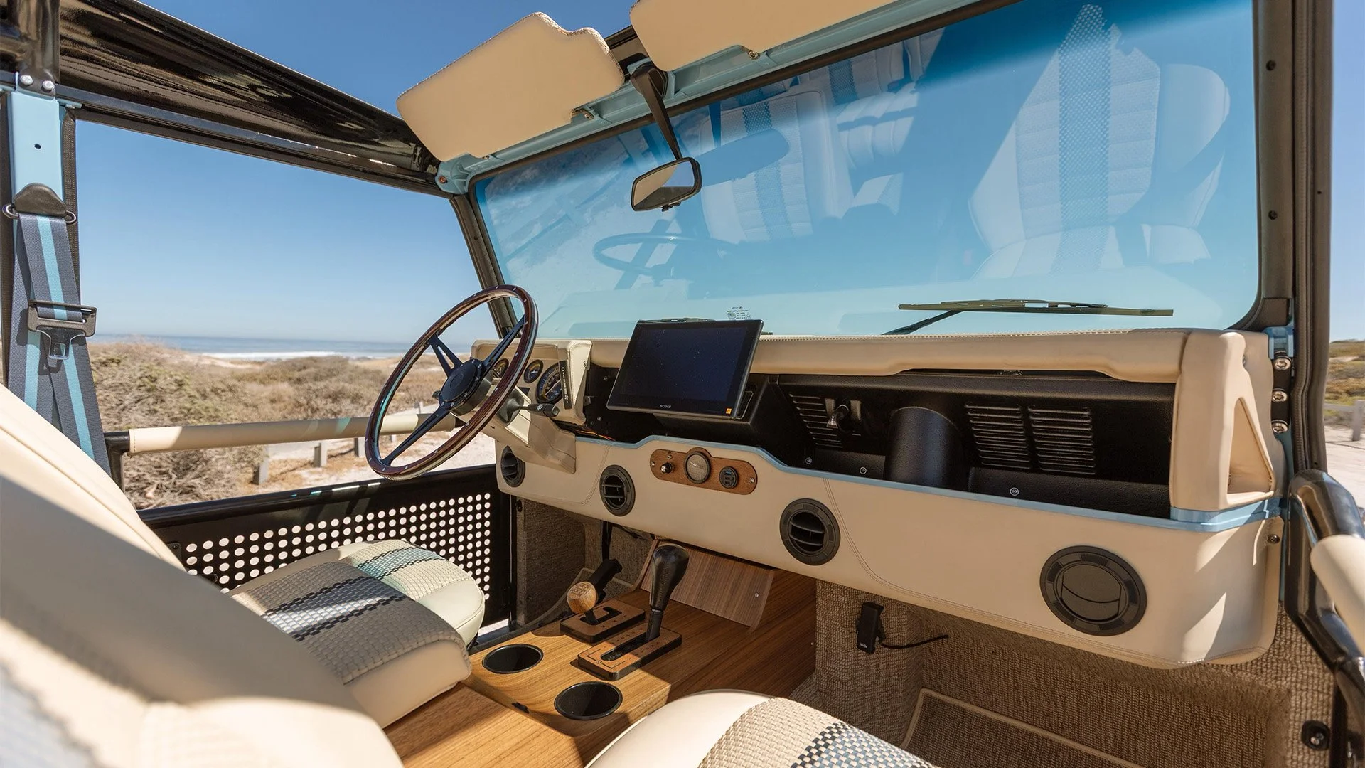 Interior of a vintage off-road vehicle parked near a beach, with a dashboard, steering wheel, and a GPS screen visible, surrounded by a clear windshield and a sunny seaside background.