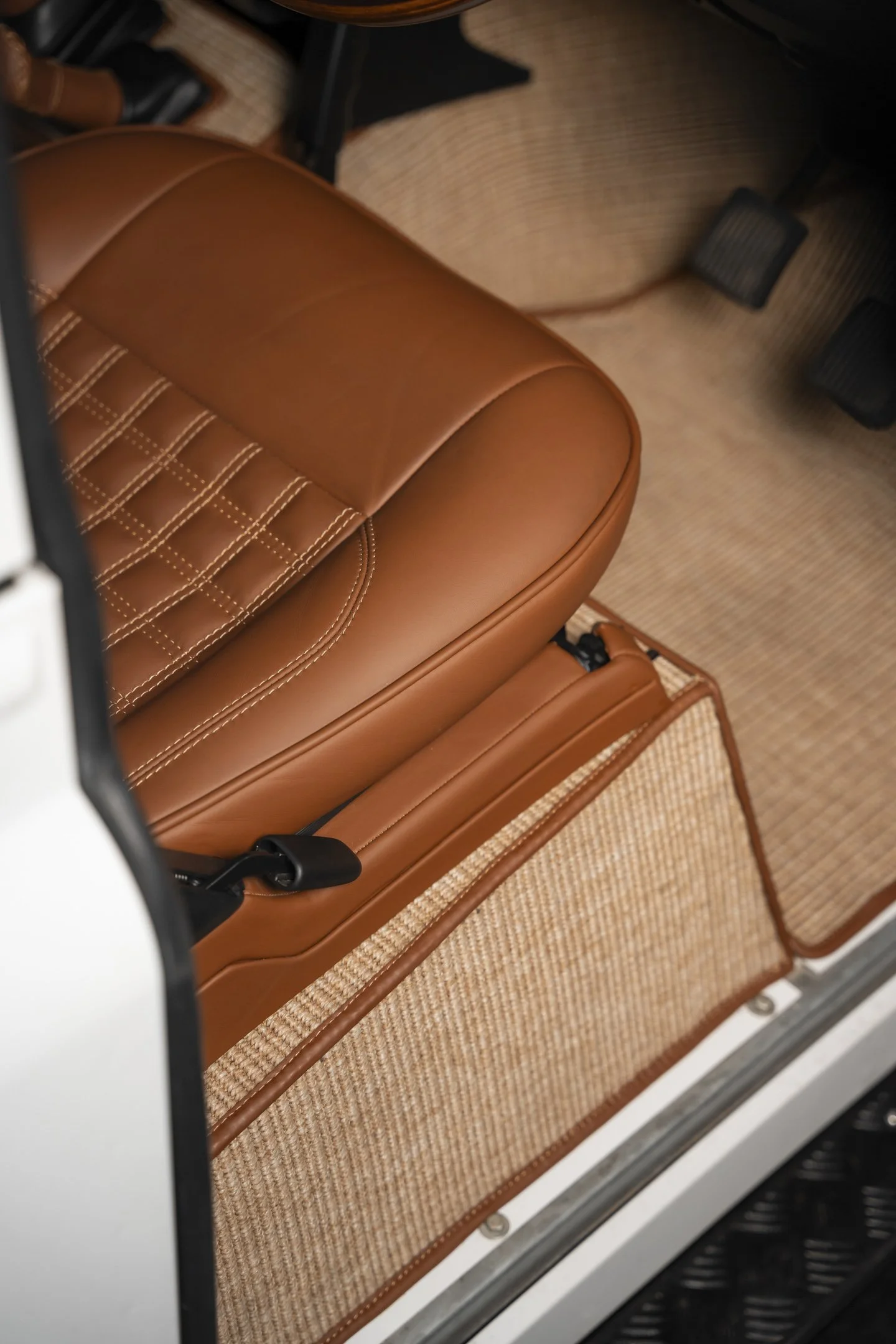 Close-up of a tan leather car seat with detailed stitching, showing the textured fabric of the floor mat and black pedals in the background.