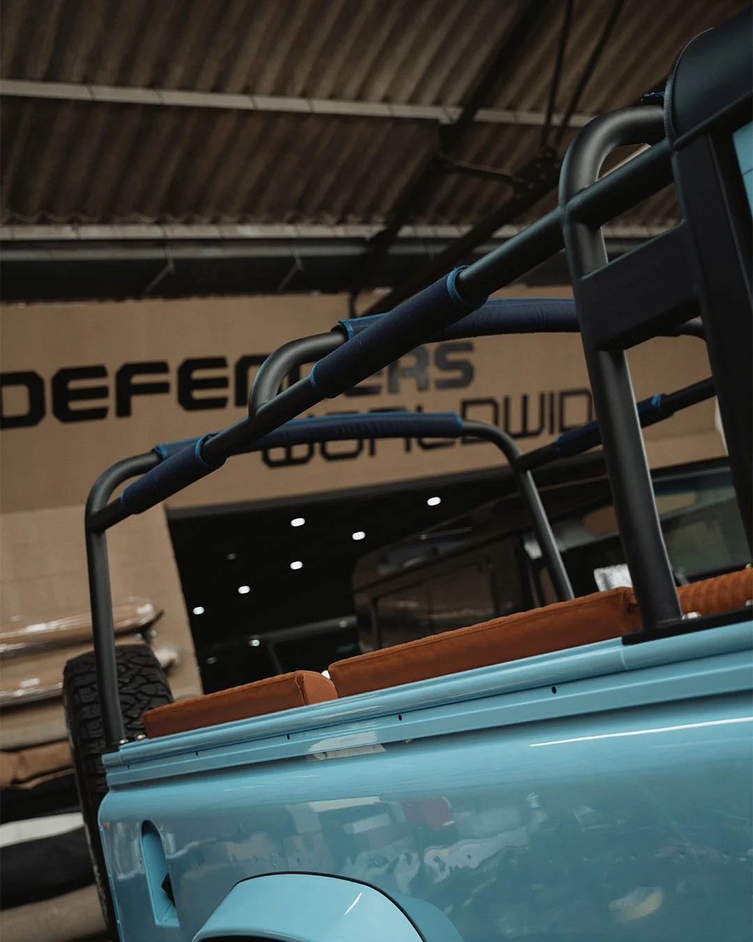 Close-up of a light blue vintage utility truck with brown leather seats in an indoor showroom with a sign that says "DEFENDERS" in the background.