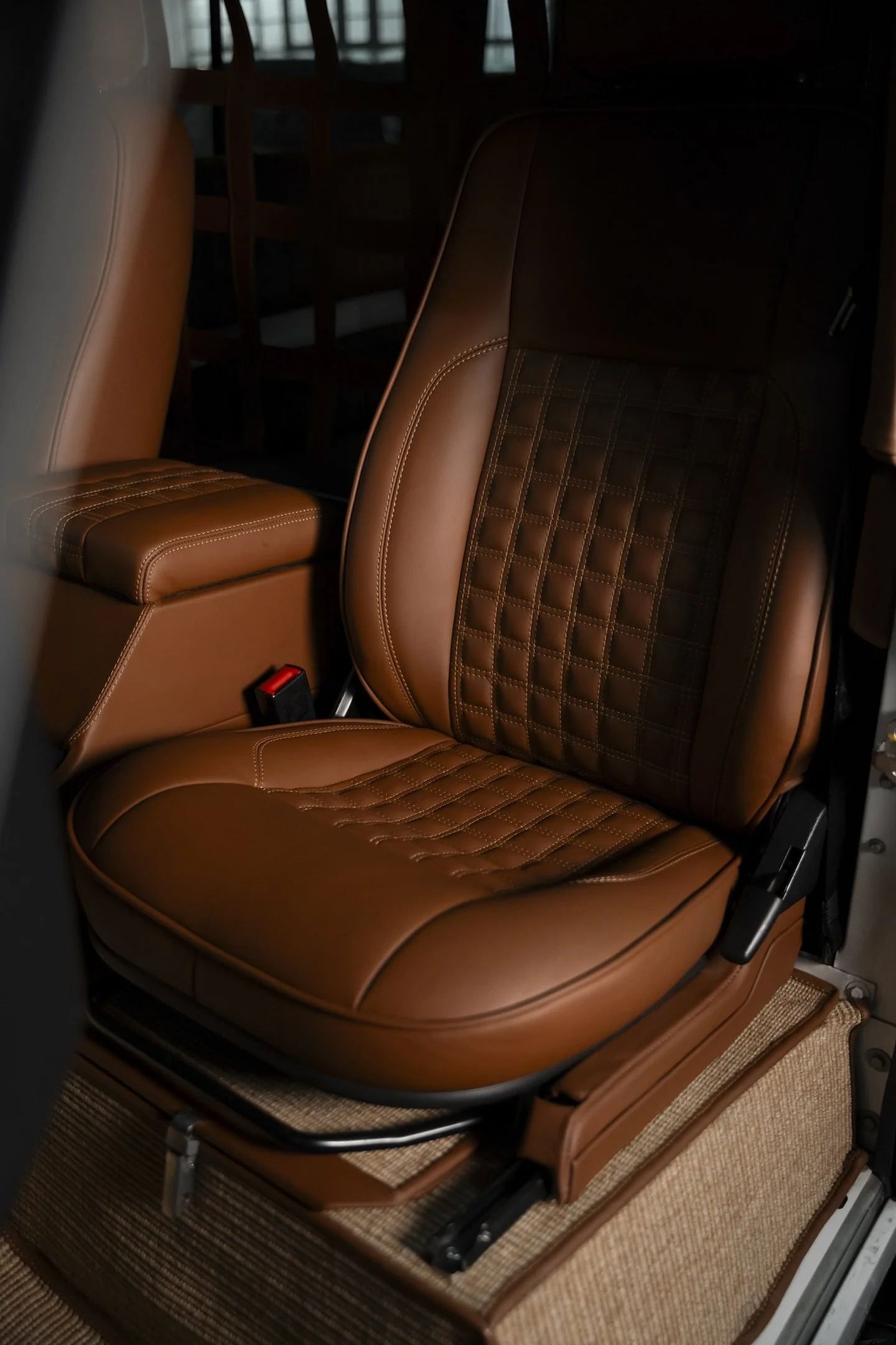 Close-up of a tan leather car seat with diamond-stitched backrest and cushioned seat, and part of a matching armrest and seat belt buckle.