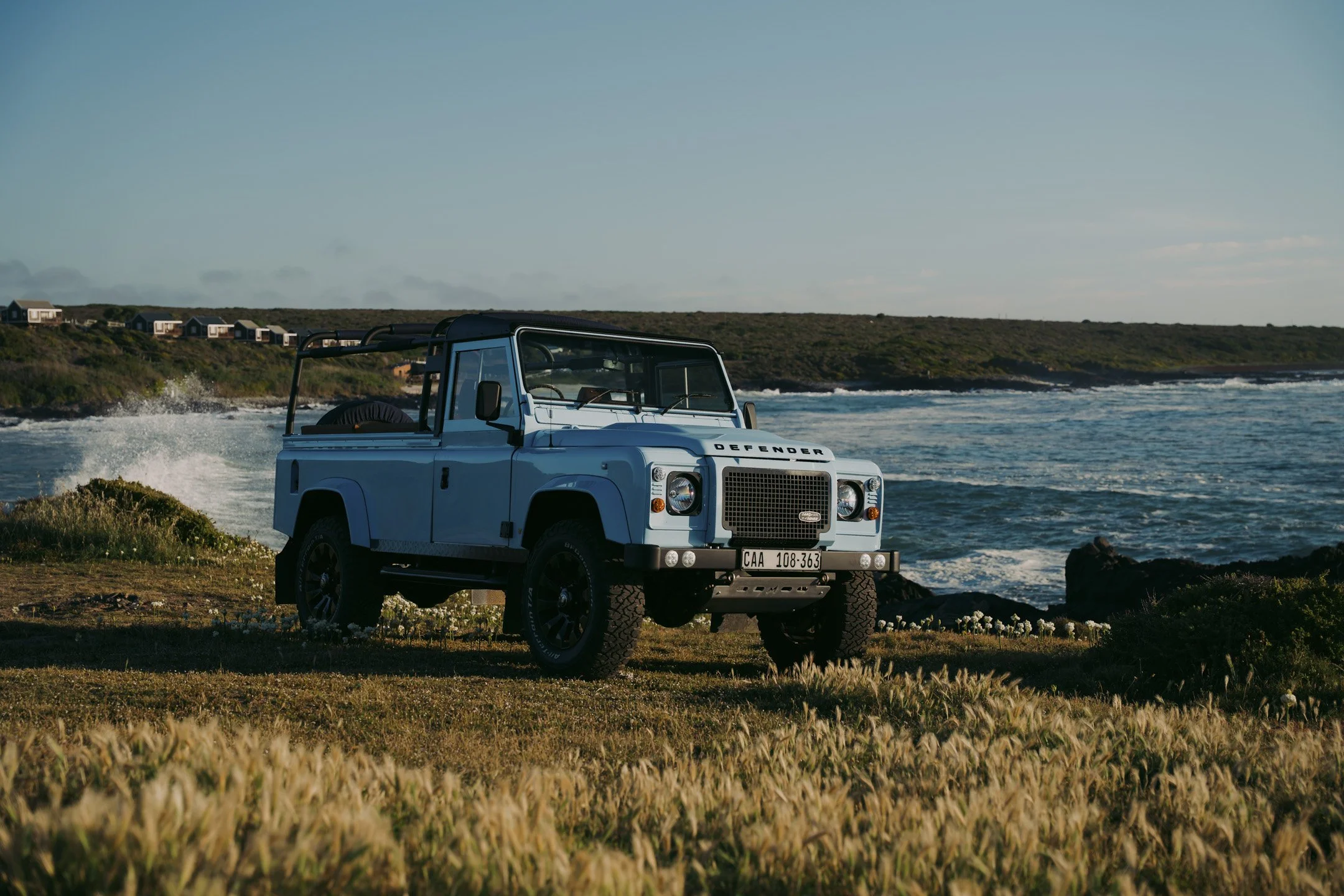 A light blue Land Rover Defender parked on grassy ground near the coast, with ocean waves and a line of beach houses on a hill in the background.