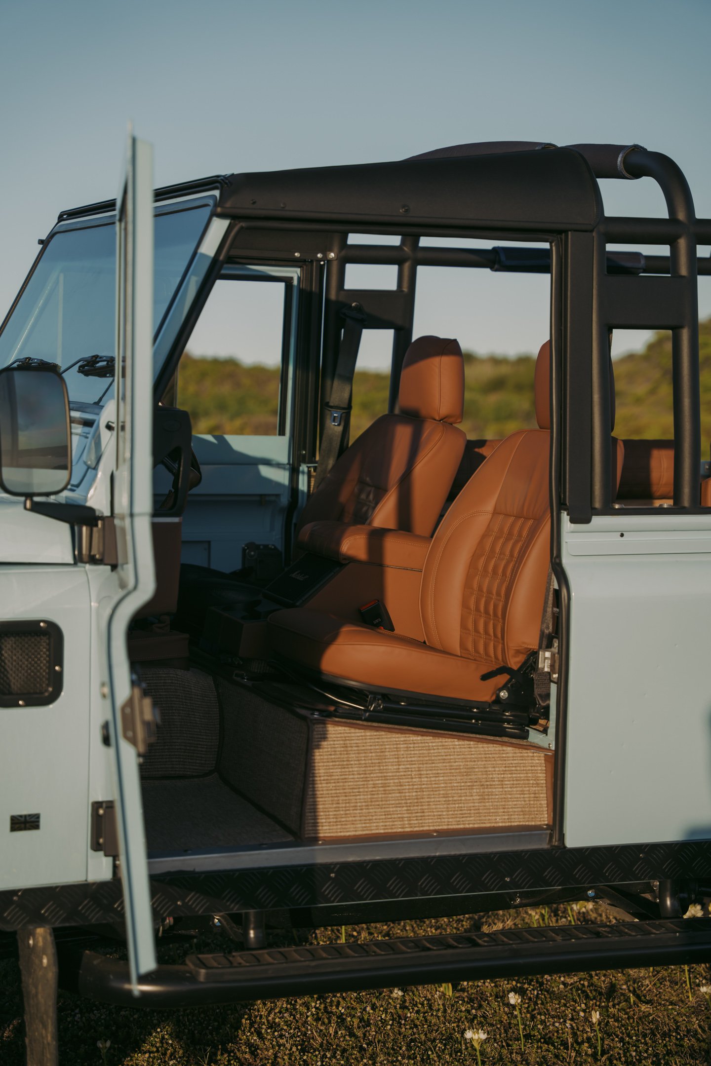 Close-up of an open vintage off-road vehicle door showing tan leather seats and black roll cage, set against grassy terrain and blue sky.