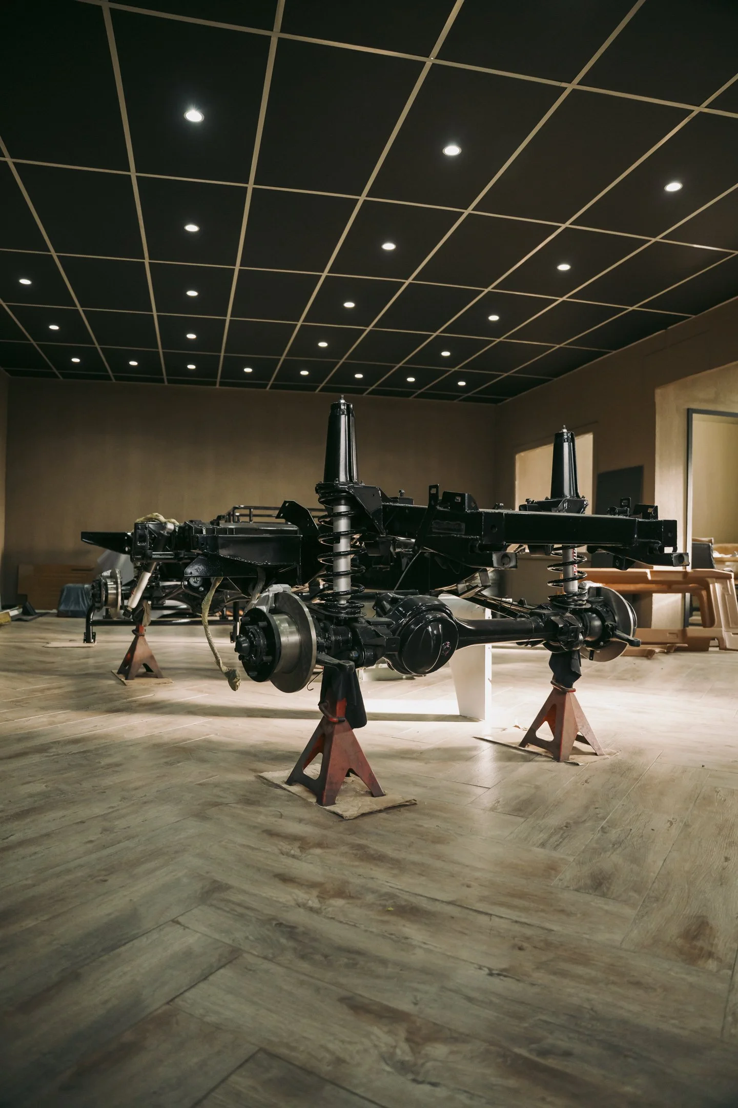 The image shows a disassembled vehicle chassis, possibly an off-road or Land Rover Defender, set on orange jack stands in a room with wood flooring and a dark ceiling with grid-like lighting.