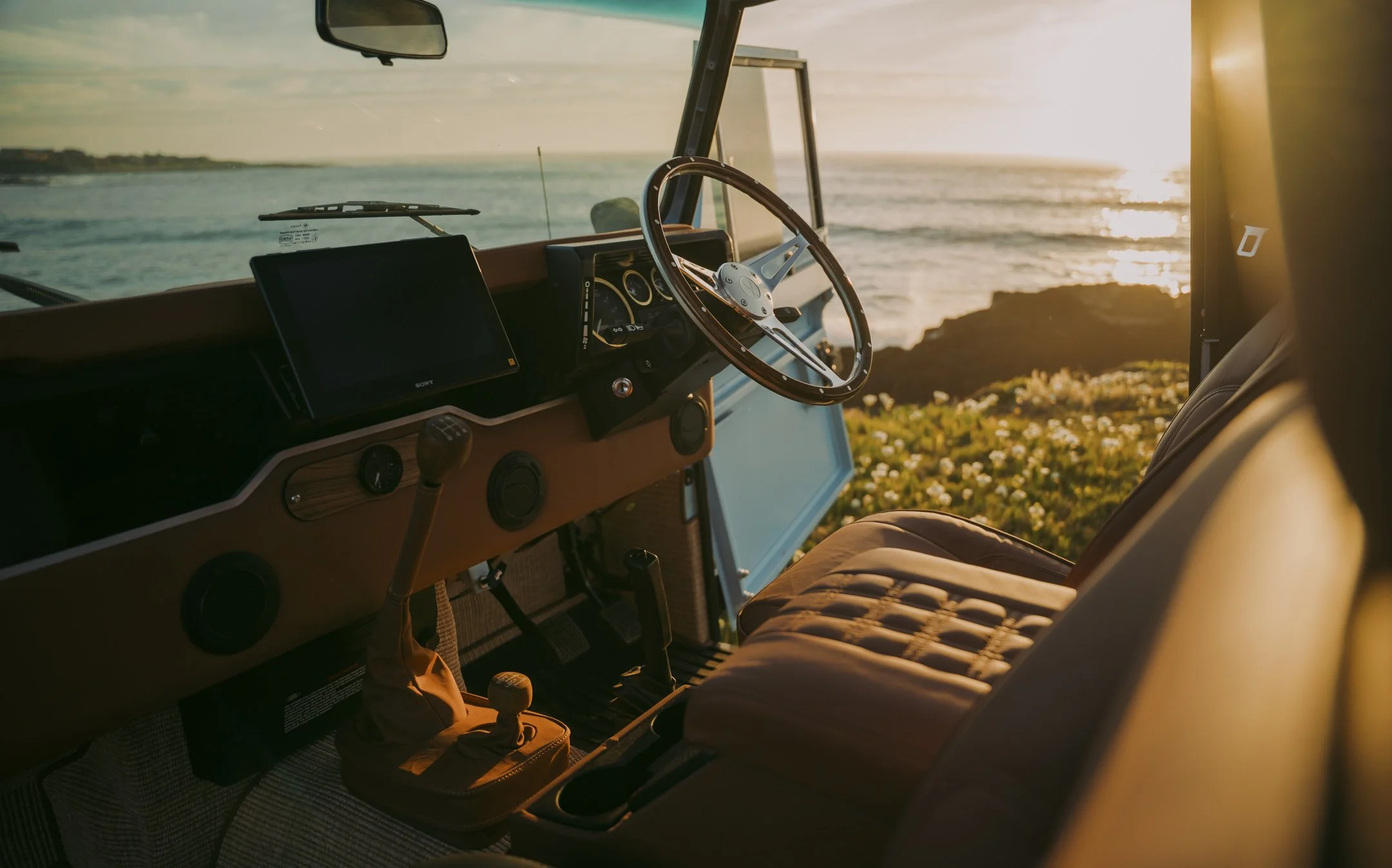 View from inside a vehicle, showing the dashboard, steering wheel, and front passenger seat with a scenic ocean and sunset view through the windshield.