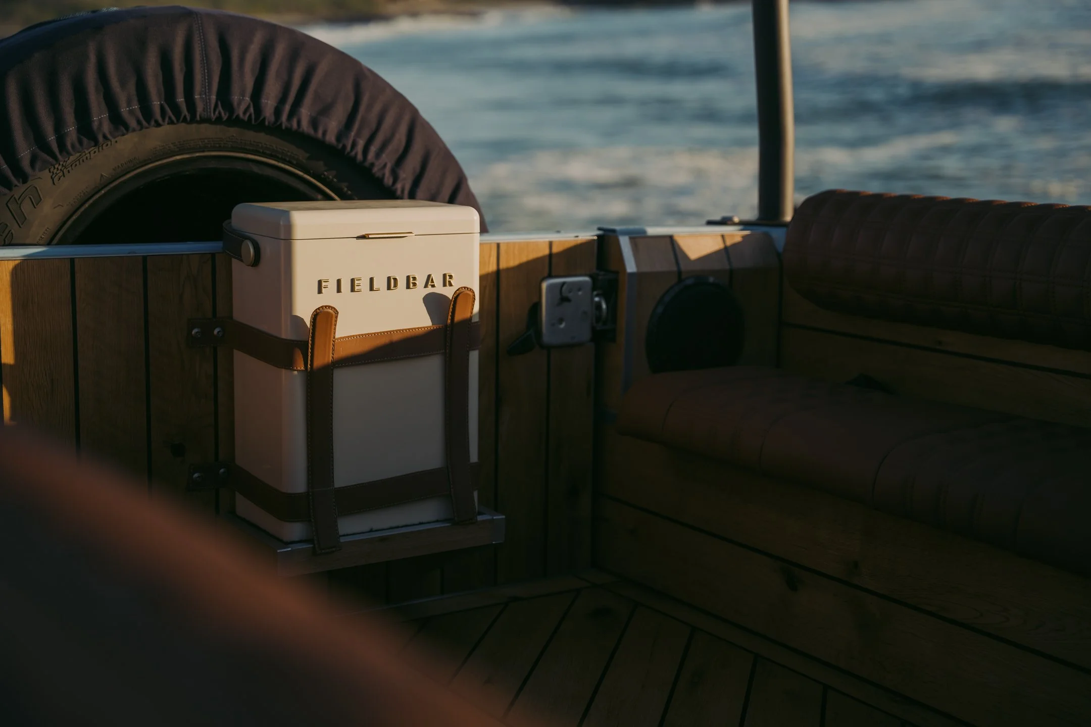 A boat's deck with a cooler labeled "FIELD BAR" secured with leather straps, a spare tire covered with a dark material, and a cushion or seating area, with water in the background.