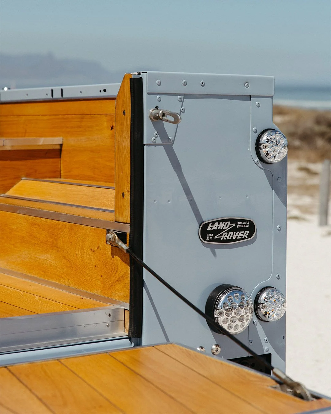 Close-up of a vintage Land Rover vehicle with wooden interior panels and a metal exterior, parked on a beach with sand and ocean in the background.
