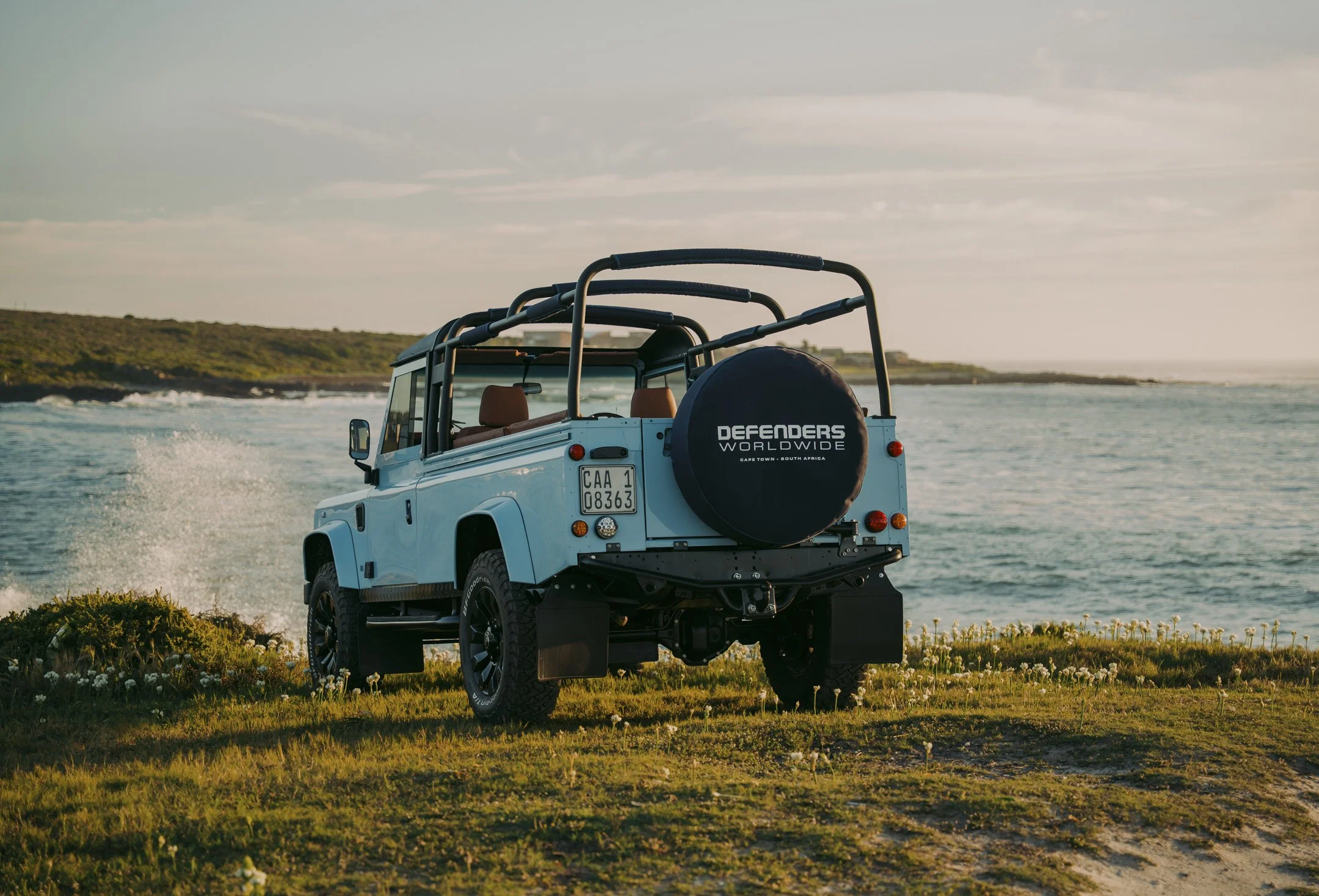 A light blue open-top off-road vehicle with a spare tire on the back, parked on a grassy patch near the beach during sunset.