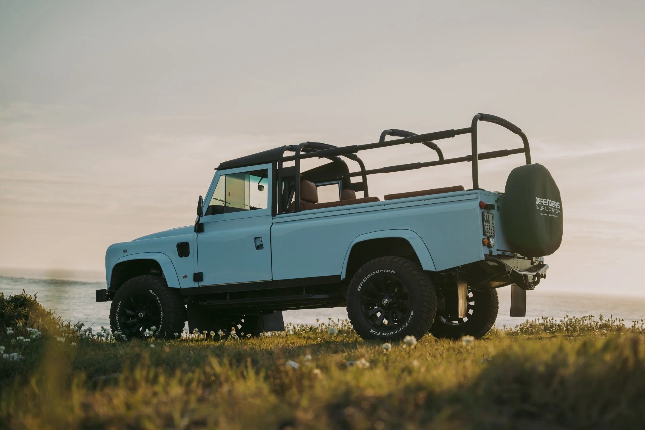A light blue off-road vehicle parked on a grassy area near the ocean during sunset, with a spare tire mounted on the back.