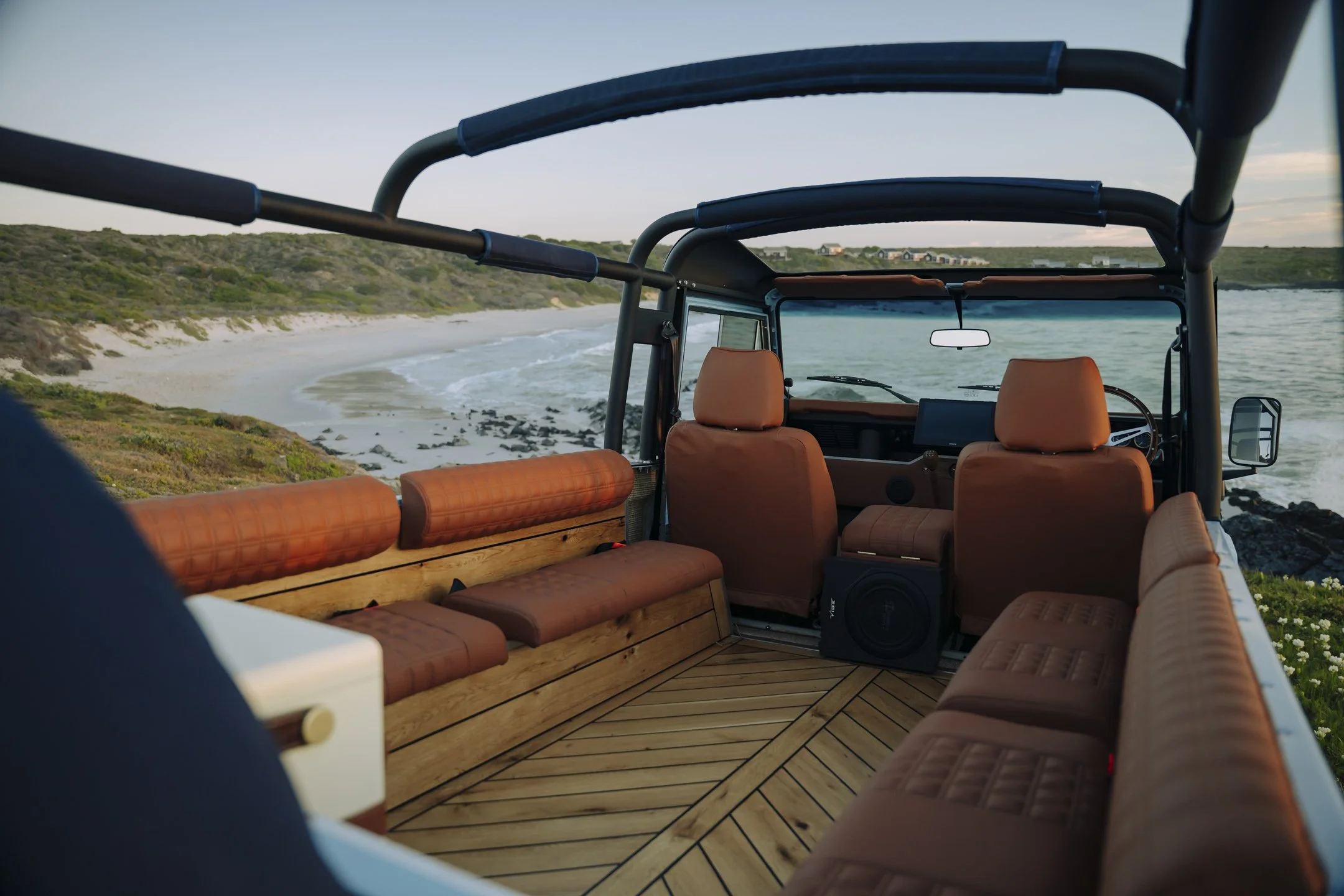 View from inside a vehicle showing a boat's interior with brown cushioned seats and a wooden floor, overlooking a beach, ocean, and grassy hills at sunset.