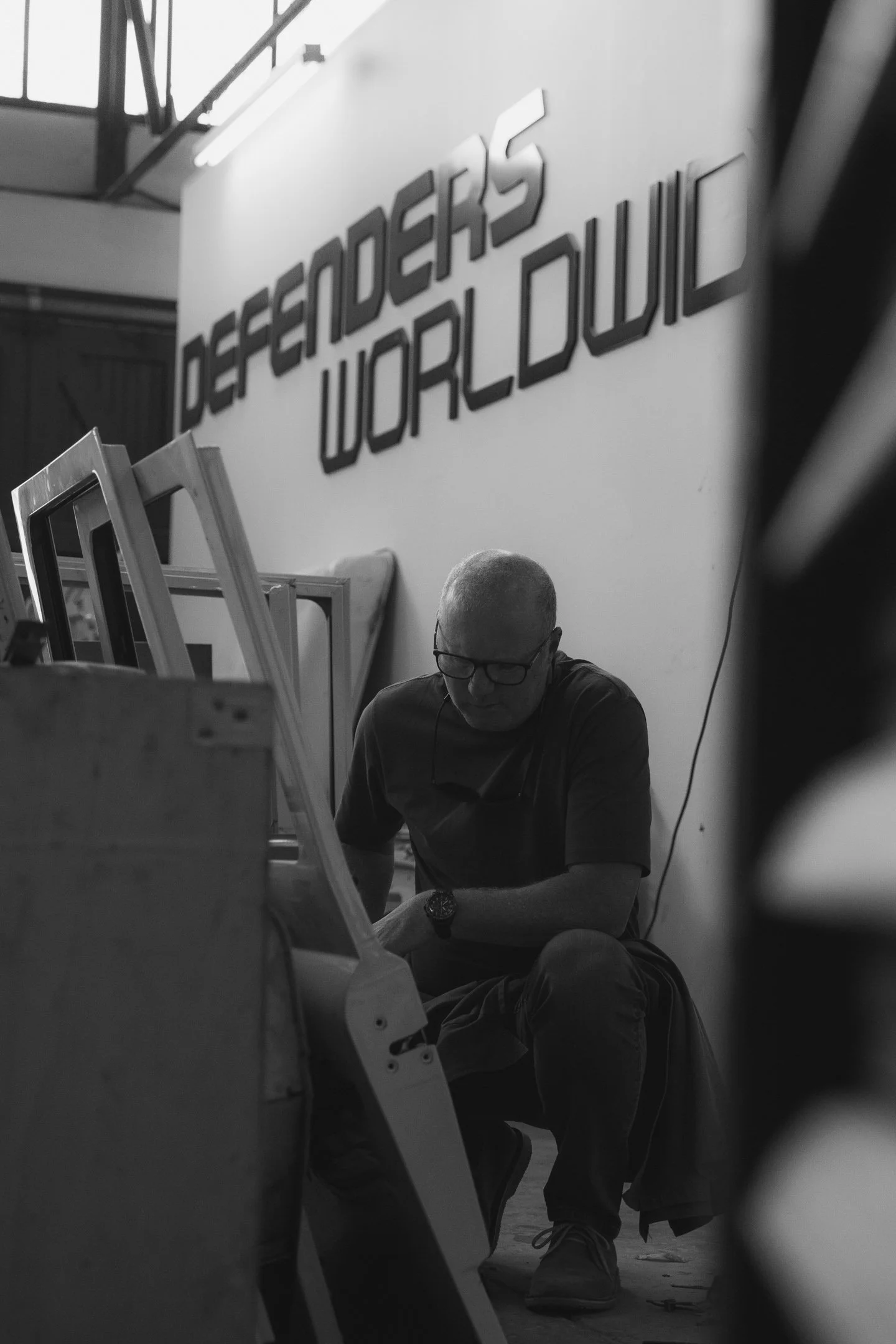 A person sitting on the floor, wearing glasses and a watch, in front of a large sign that reads 'DEFENDERS WORLDWIDE' in a building with industrial design elements.