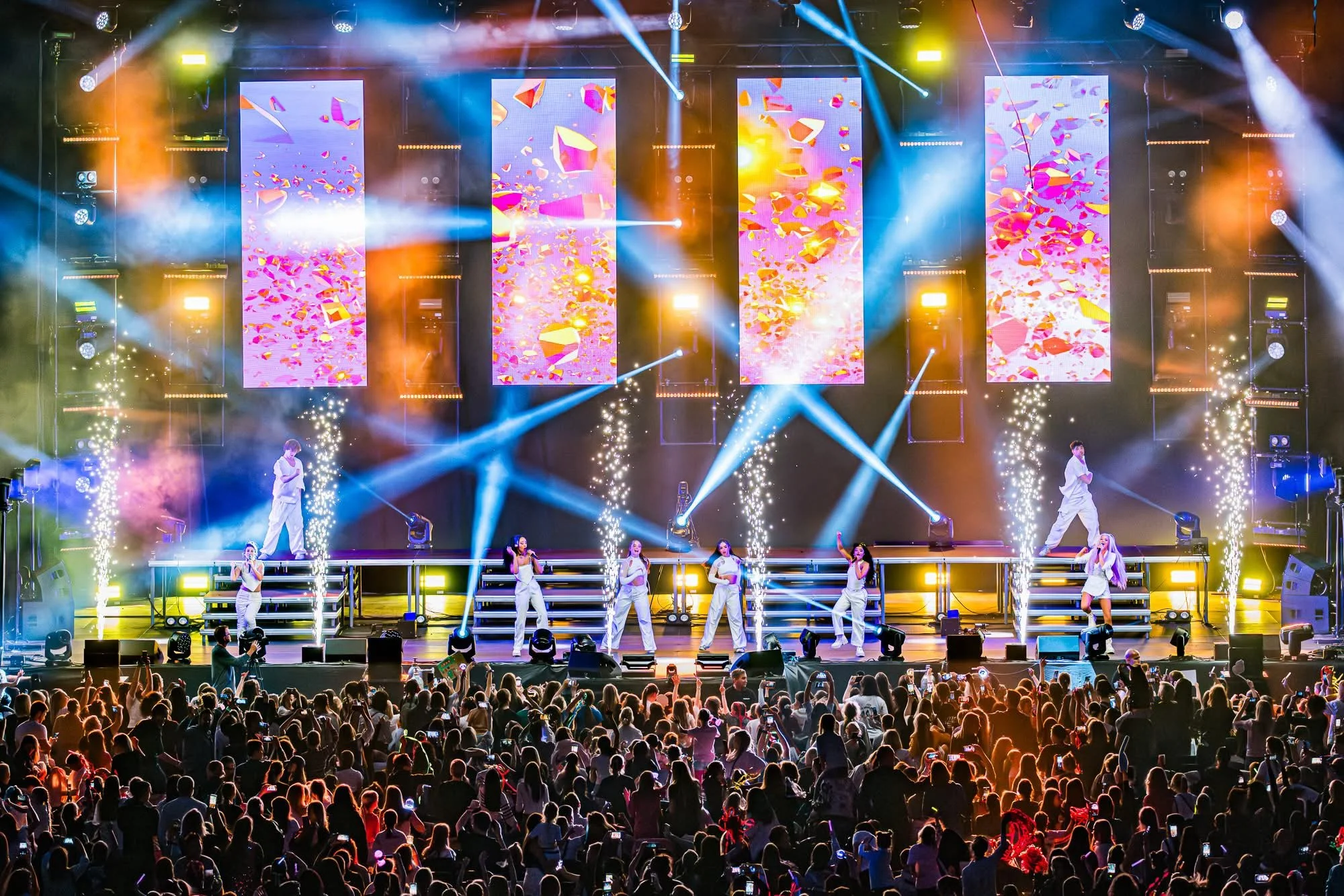 A lively concert stage with performers, colorful large LED screens displaying abstract designs, vibrant lighting effects including beams and spotlights, and audience members watching and taking photos in a large indoor venue.