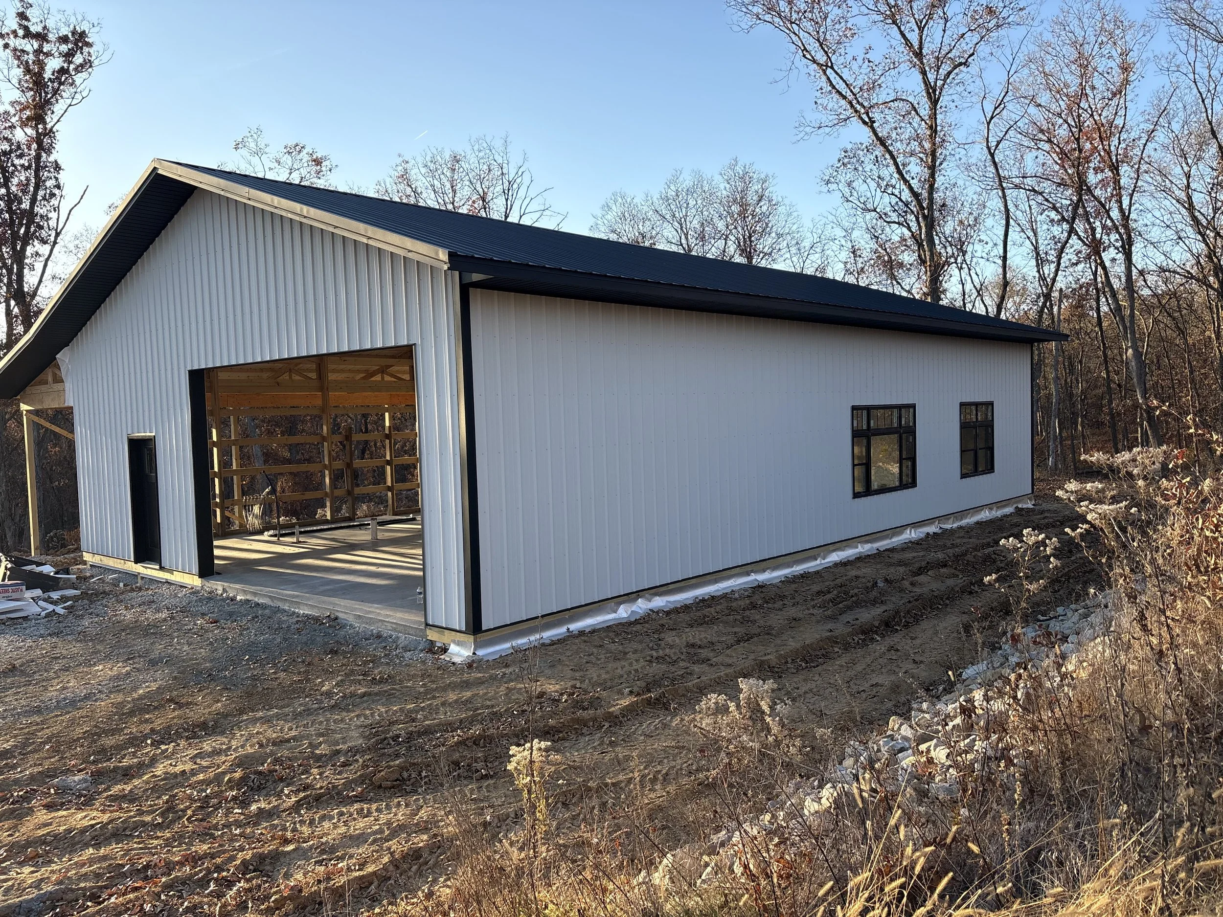 A pole barn under construction with white metal siding on the walls and a black metal roof, located in a wooded area with trees and clear sky in the background.