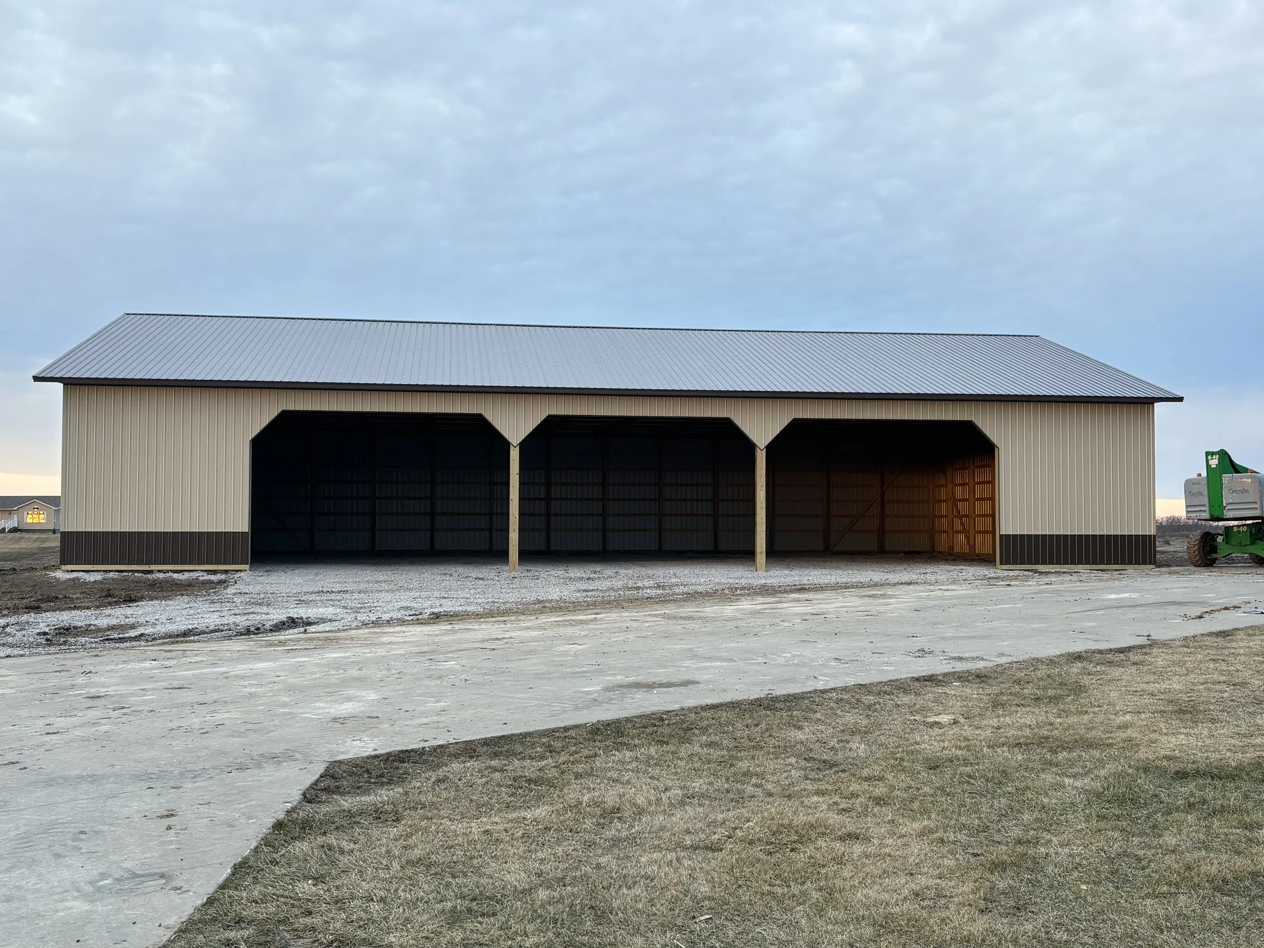 A large, open metal shed with a gabled roof, beige walls, and large open doors, situated outdoors on a gravel and grassy area under a cloudy sky.