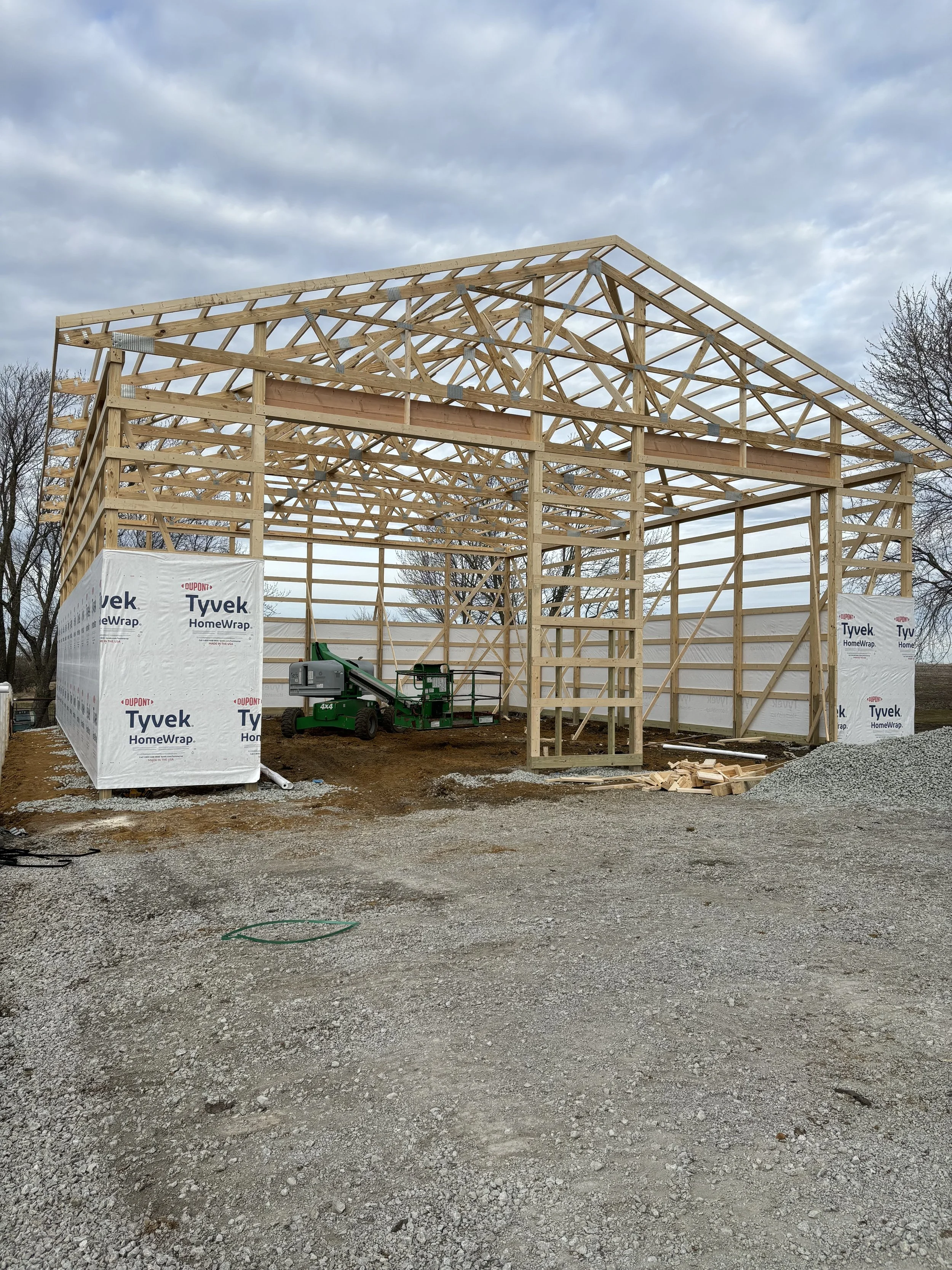 Construction site with new wooden framework for a building, wrapped in Tyvek HomeWrap, with construction equipment and gravel ground, and cloudy sky overhead.