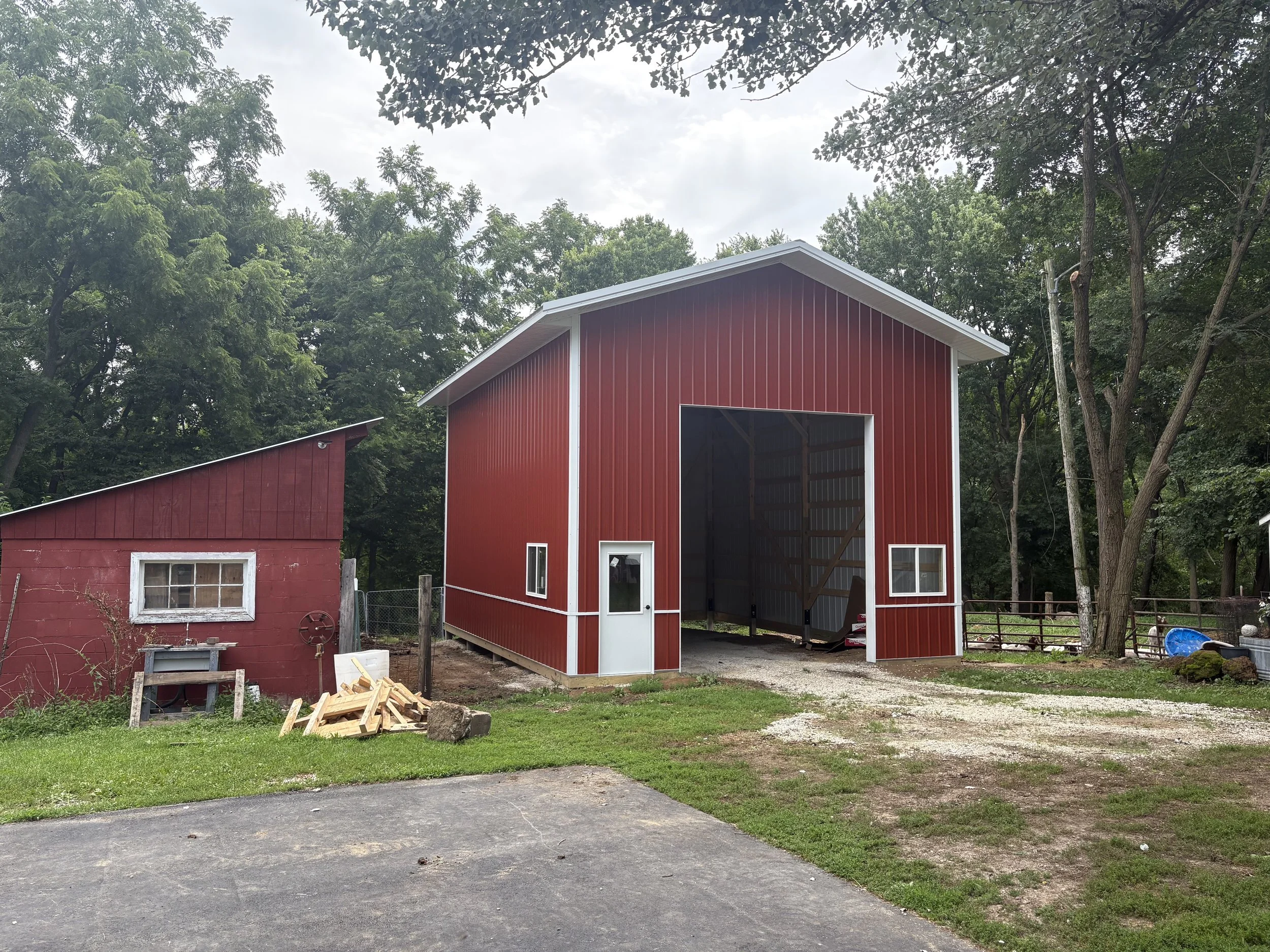A red barn with white trim, including a large open doorway and a small window on the front. Next to the barn, there is a smaller red shed with a window. The area around the buildings features grass, dirt, and gravel with scattered wood and farm equip