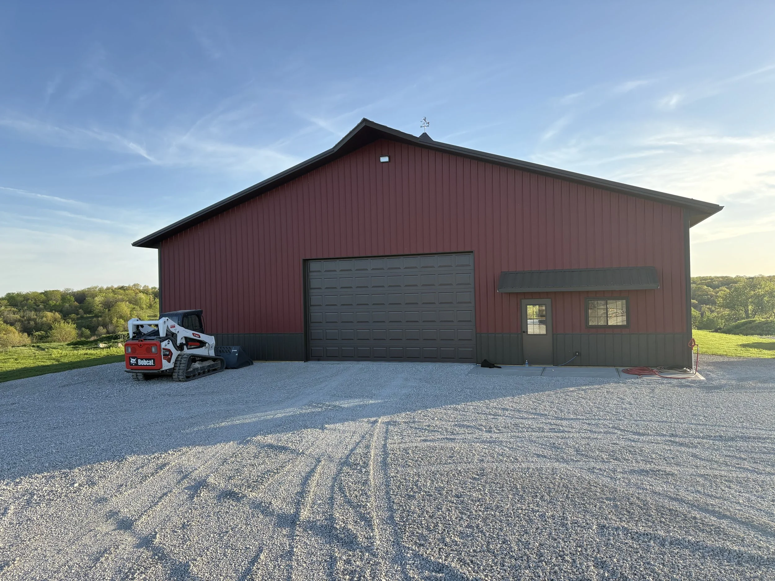 A red barn with a gray garage door, a small window, and a black awning. There is a Bobcat skid-steer loader parked on a gravel driveway in front of the barn. The sky is clear with wispy clouds, and there are green trees and hills in the background.