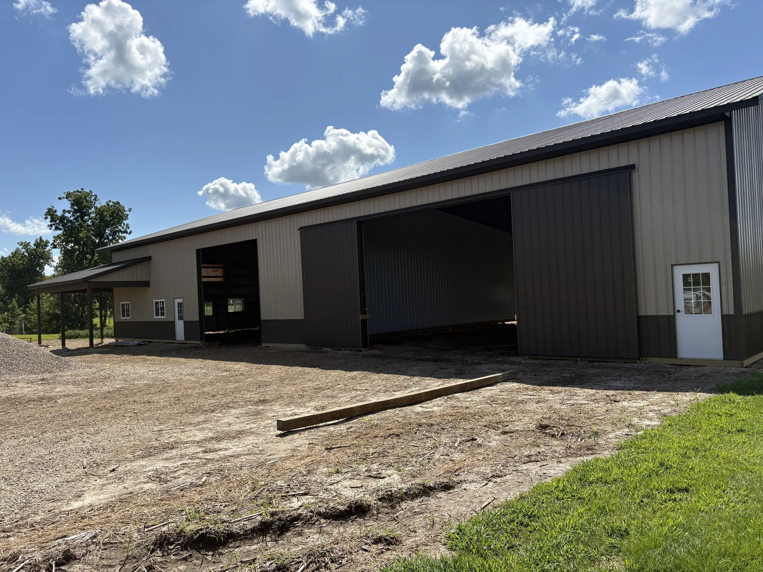 A pole building with large open doors, set in a rural area with trees and a partly cloudy sky.