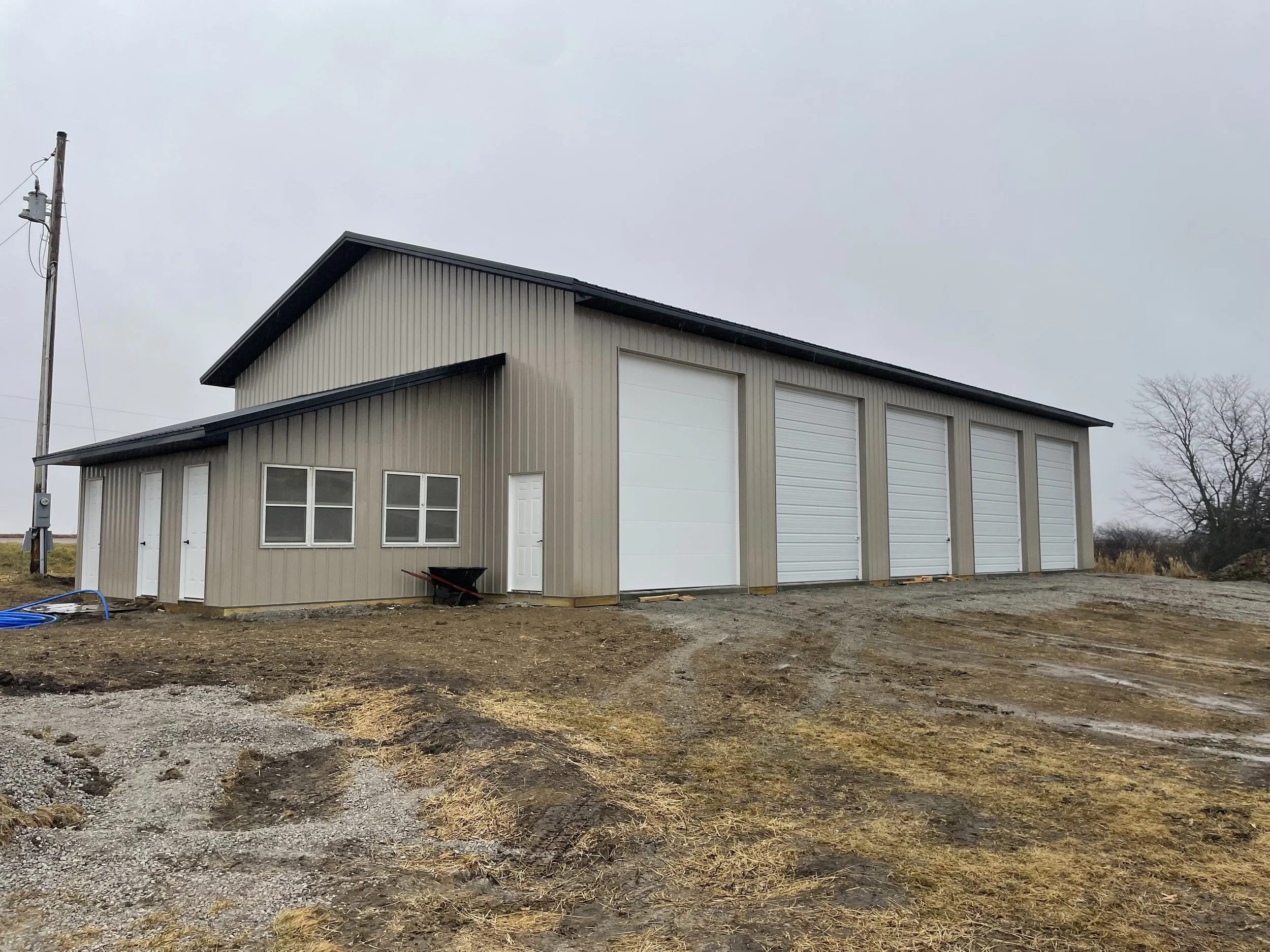 A large modern barn with a metal exterior, multiple large white garage doors, a small entrance door, and a sloped roof. The barn is situated in a rural area with a grey, overcast sky and a few leafless trees in the background.