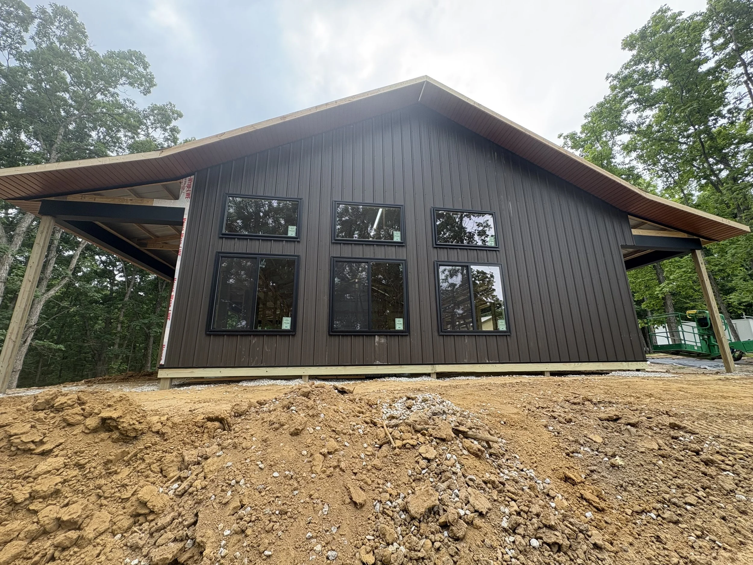A house under construction, with black vertical siding and multiple large windows, situated on a dirt lot surrounded by trees, with a partly cloudy sky overhead.