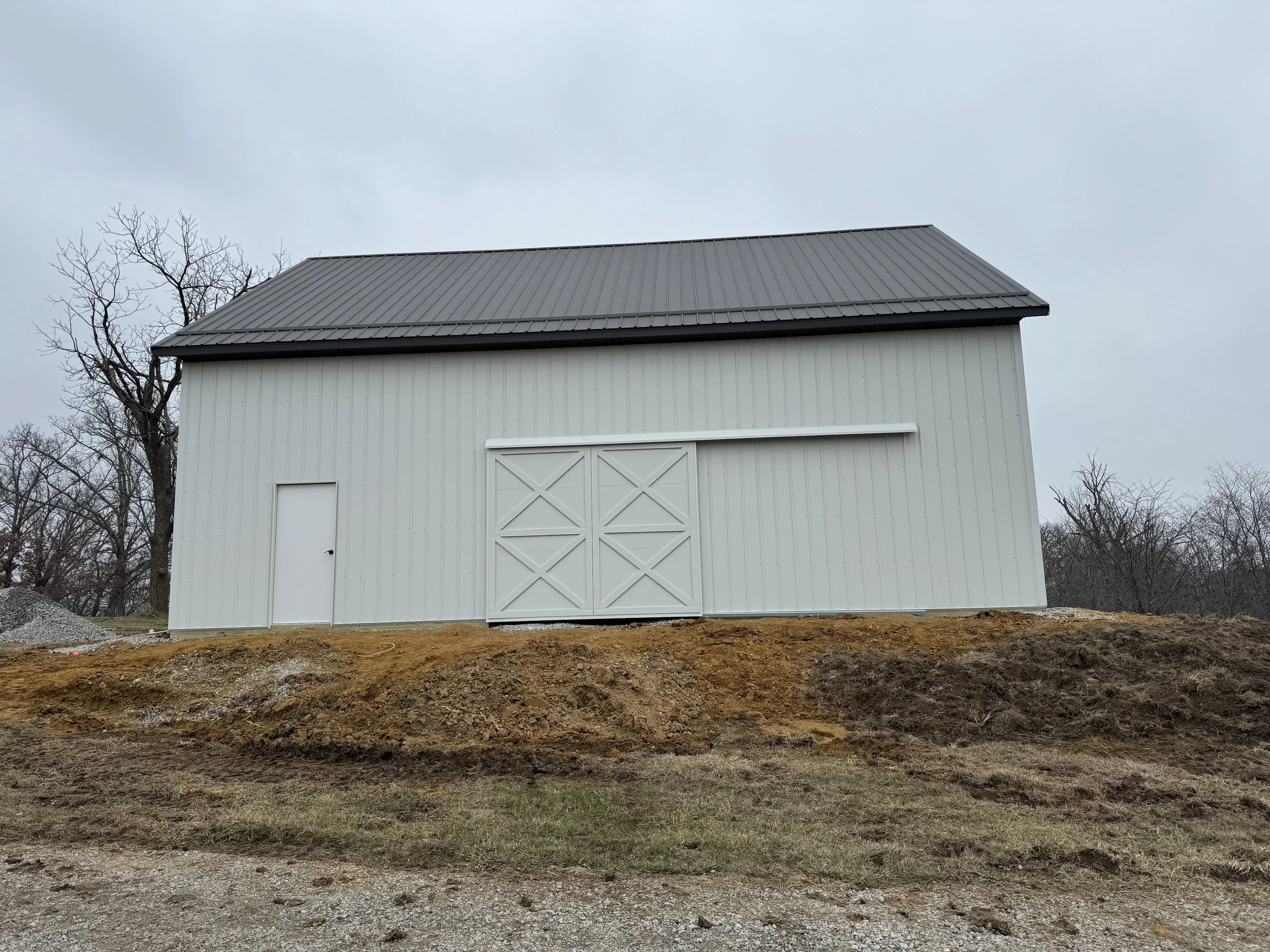 A white barn with a gray metal roof, surrounded by leafless trees and dirt ground, under a cloudy sky.