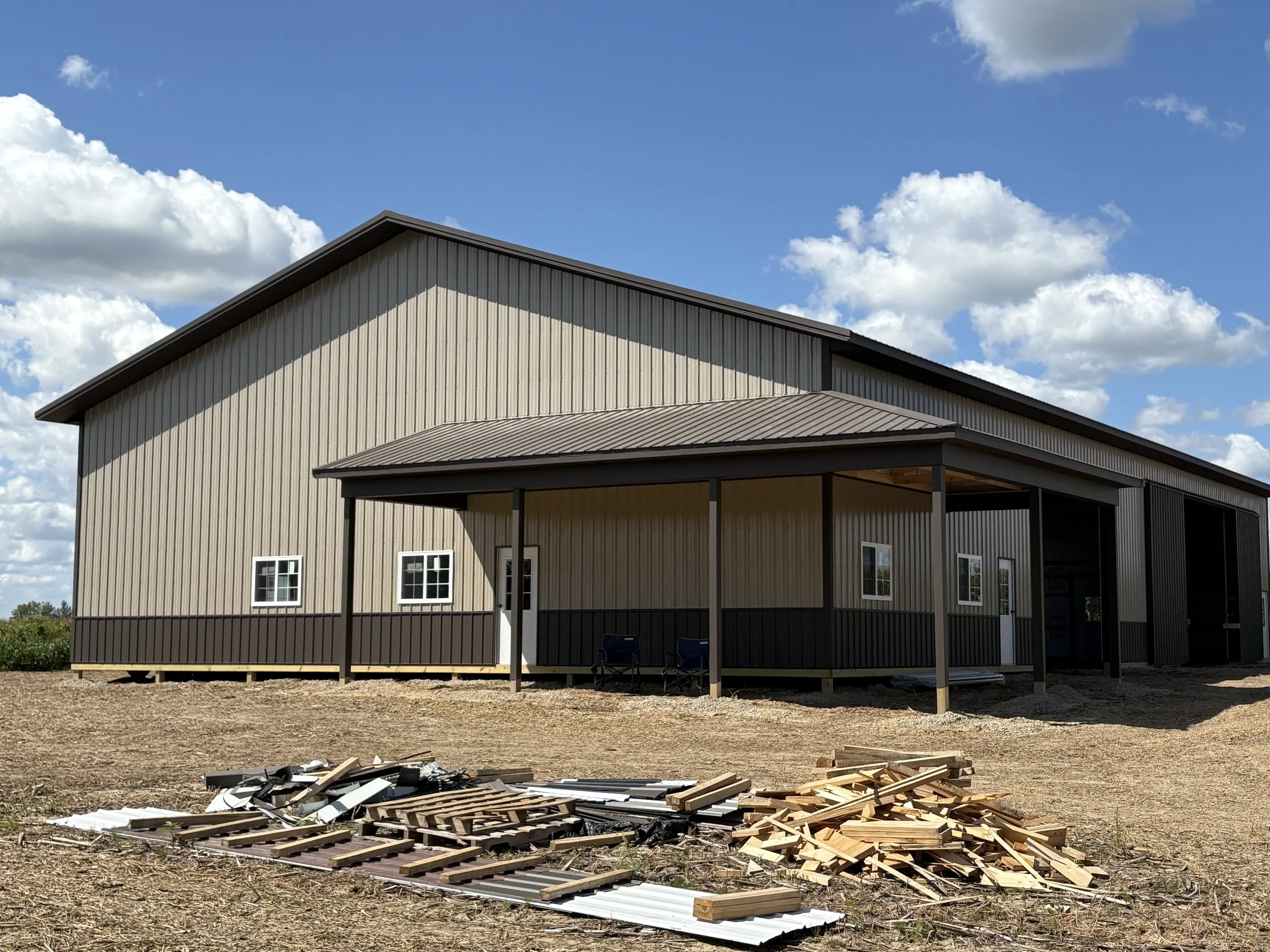 A partially constructed building with a metal roof and siding, under a blue sky with scattered white clouds. Construction debris and building materials are scattered on the ground in the foreground.