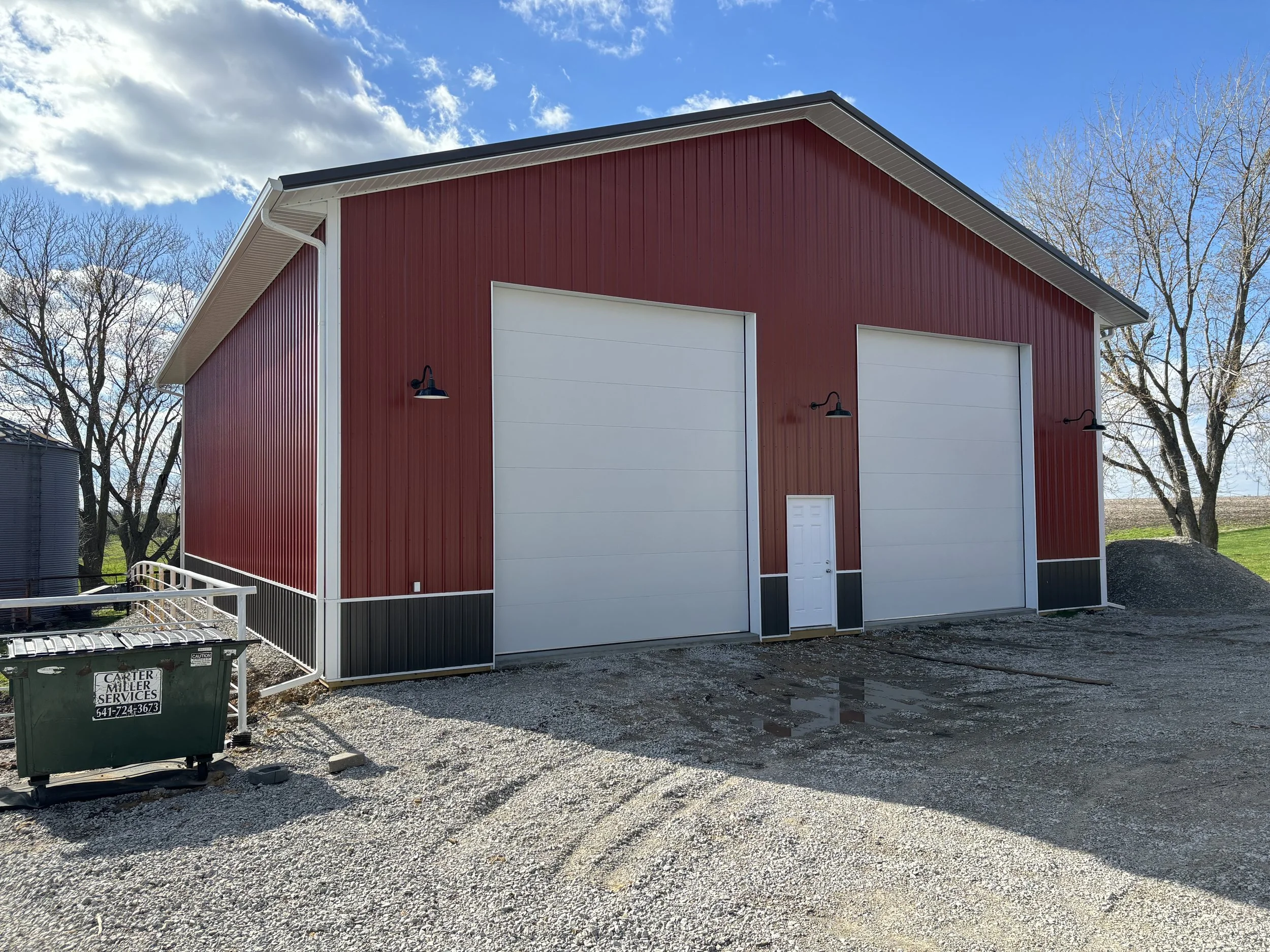 A red barn with white trim and two large white garage doors, outdoor lighting fixtures, a small door, metal railing, trash bin on gravel ground, and leafless trees in the background under a partly cloudy sky.